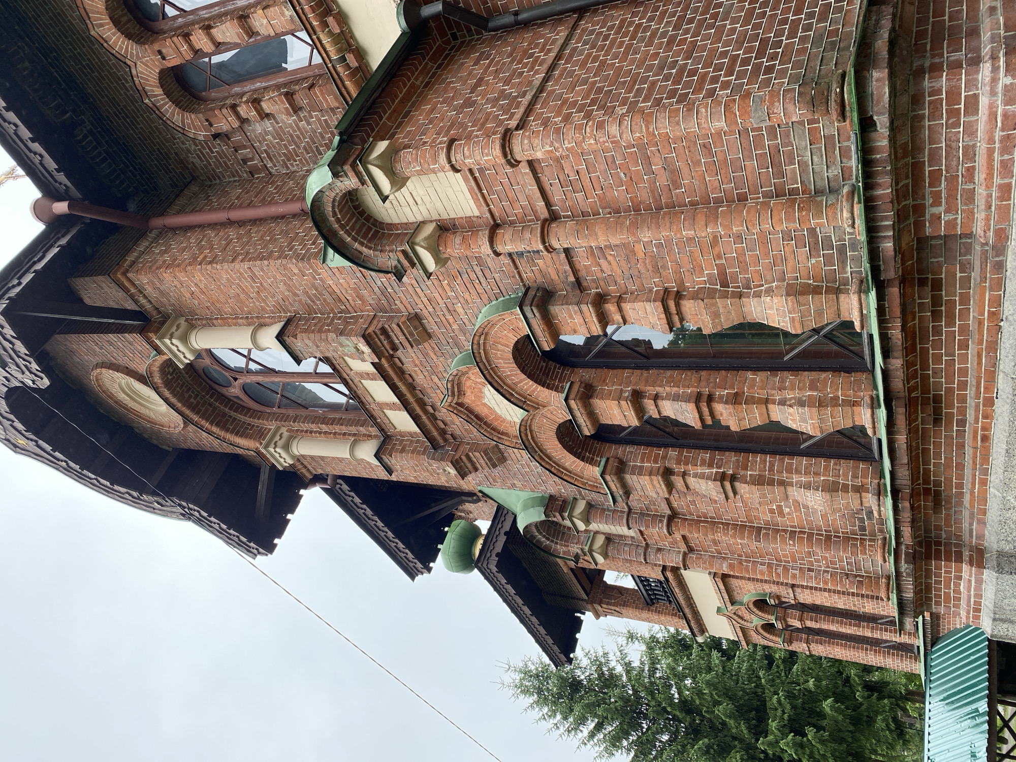 An outdoor, low-angle, full shot captures the exterior of the Valaam Monastery, located in Priozersk, Russia. The building is constructed of red brick with decorative white trim and green accents. Arched windows, some with multiple panes, are visible. Wooden eaves with decorative trim overhang the building. A small green dome peeks out from behind the eaves on the left side. A tall evergreen tree is visible to the left of the building. A portion of a green metal roof structure is in the foreground at the bottom left of the image. The sky is overcast and a single wire runs diagonally across the upper portion of the frame.