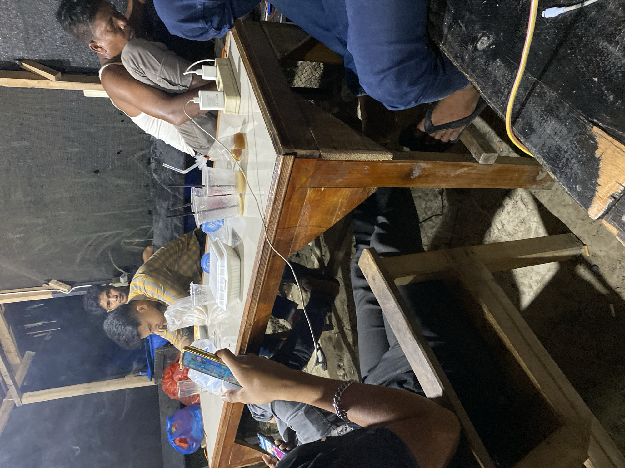 The image captures a casual gathering of several individuals, primarily men, around a wooden table in what appears to be a dimly lit, informal indoor or sheltered setting, likely at night. The scene features a mix of socialization and individual engagement with mobile devices.
Around the wooden table, several objects are visible, including clear plastic cups with straws, some containing beverages, and transparent plastic bags possibly holding food items. A white power strip with multiple charging blocks and cables indicates active device charging. White plastic containers are also present on the tabletop.
At least five people are gathered. One man, wearing a white tank top and light-colored shorts, is seated with his back against a dark wall, looking towards the table. Another individual in blue jeans and a dark top is visible, with their foot, adorned with a black flip-flop, resting on the ground. Towards the left side of the table, a man in a plaid shirt is seen looking down, while another man is partially visible behind him. In the immediate foreground, a person wearing a black top and a silver chain bracelet is engrossed in a smartphone, with another phone visible nearby.
The ground beneath the table appears to be uneven, possibly dirt or rough concrete. Wooden beams are part of the surrounding structure, suggesting a simple or makeshift shelter. A light cloud of smoke drifts in the background on the left, hinting at a nearby cooking fire or heat source. The atmosphere is relaxed, with people appearing to be unwinding and interacting with their phones. Faint text "CE" and "20W" are visible on one of the white charging blocks on the table.