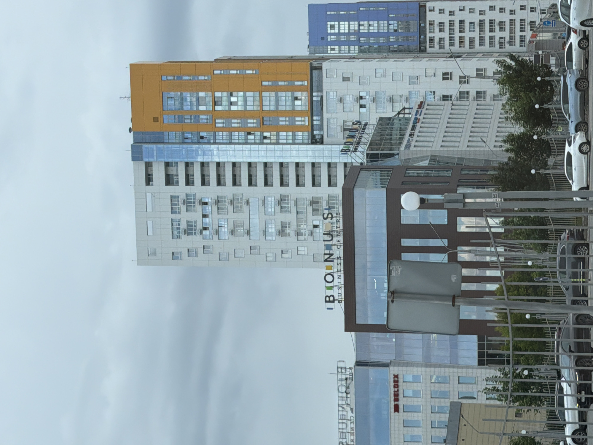 The image shows a street view of modern high-rise buildings in the city of Perm, Russia. The buildings are of various architectural styles and colors, including white, orange, blue, and brown. Some of the buildings have large glass facades, reflecting the cloudy sky. One building prominently displays a sign that reads "BONUS BUSINESS CENTRE". The foreground features a metal fence, streetlights, and several cars parked along the street. The overall impression is of a contemporary urban environment.