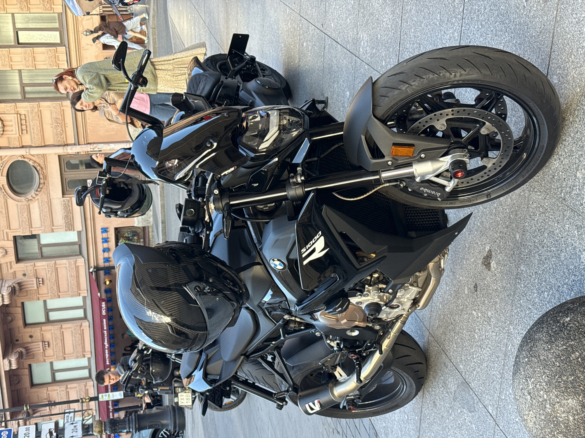 This image captures a black BMW S1000 motorcycle parked on a paved city street in Saint Petersburg, Russia. The motorcycle is in the foreground, positioned at an angle with its front wheel slightly turned to the right. A black, carbon-fiber patterned helmet rests on the seat. In the background, there are buildings with classical architecture, and several people are visible walking on the sidewalk and in the street. One person appears to be interacting with the motorcycle. The street is made of light-colored paving stones, and a large, round, stone planter is present in the lower left corner of the frame. The scene appears to be a daytime urban setting.