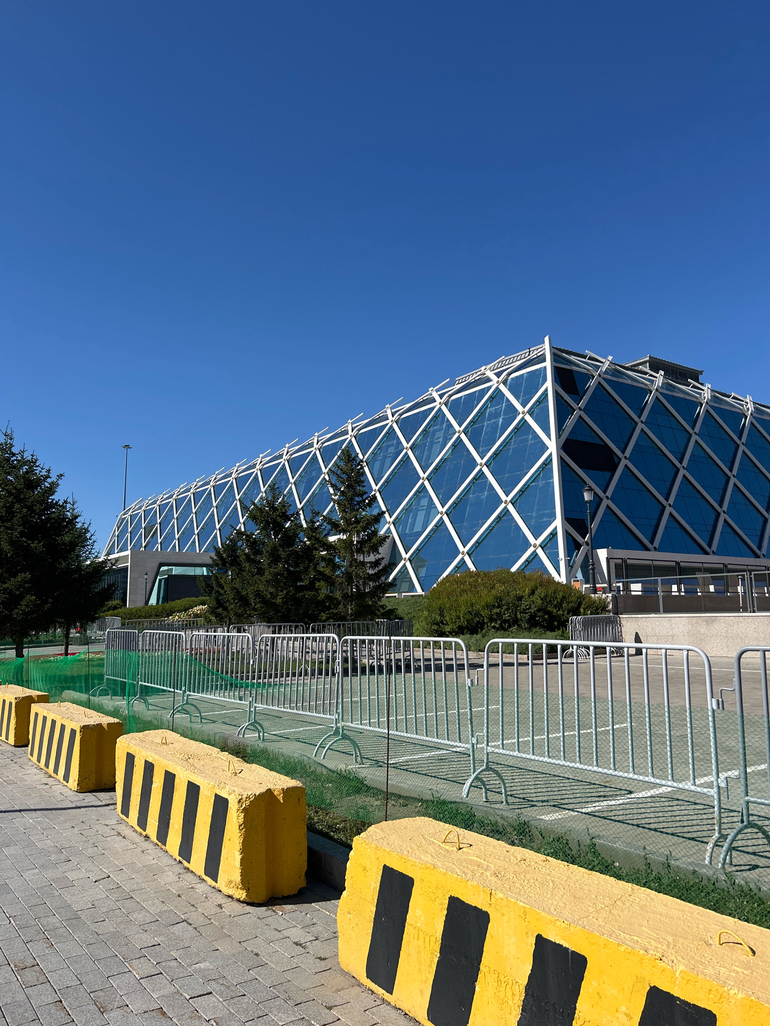 The image shows the National Pavilion of Kazakhstan at Expo 2017 in Astana, Kazakhstan, on a clear, sunny day. The pavilion is a striking geodesic dome structure covered in blue glass and a white lattice framework. In the foreground, there are several yellow concrete barriers with black diagonal stripes, placed along a paved walkway. Behind the barriers, a silver metal fence separates the walkway from a green, grassy area. Beyond the fence, trees and bushes are visible, with the pavilion forming the backdrop. The sky is a vibrant, cloudless blue. There are no people visible in the image.