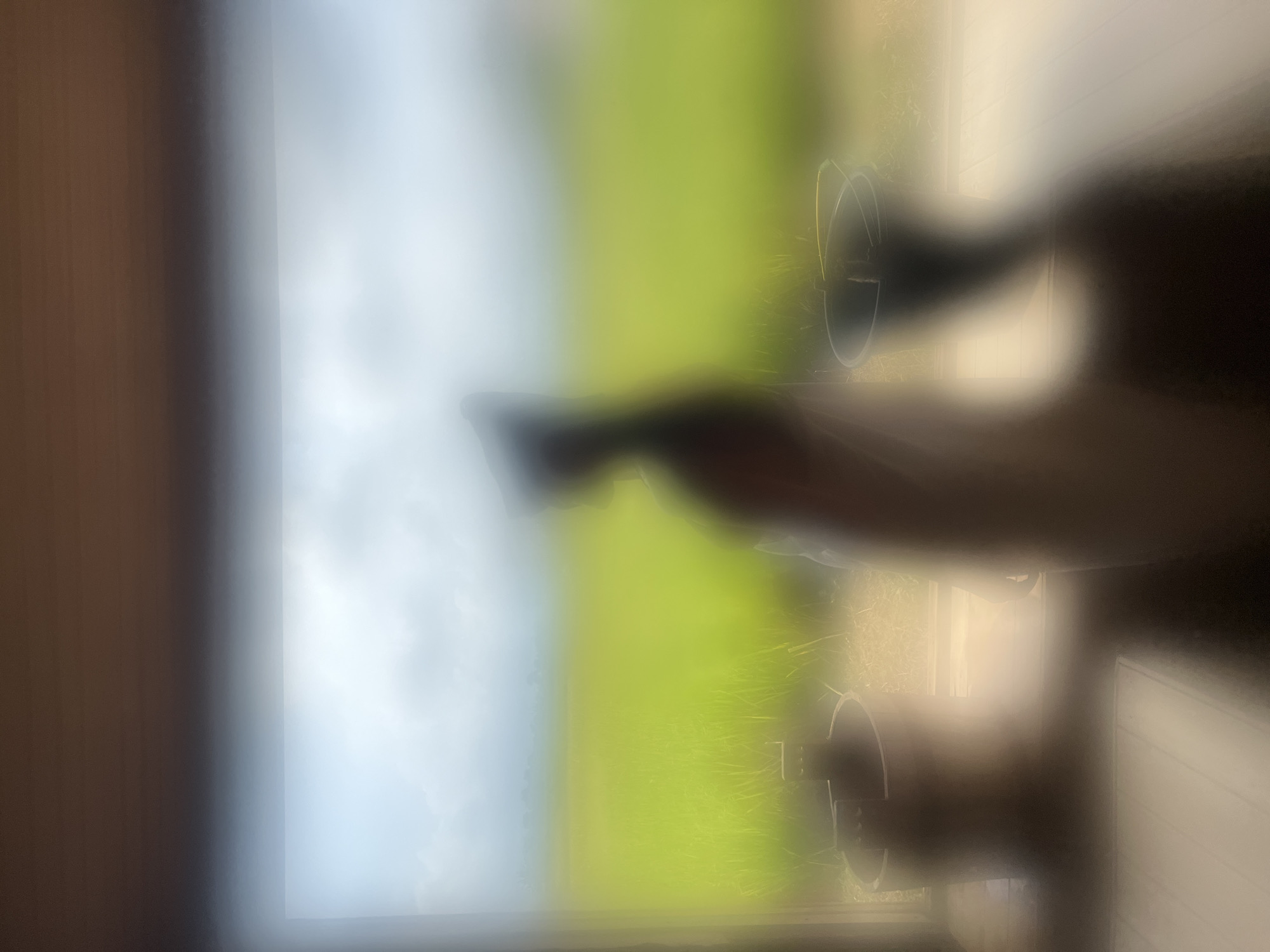 The image shows a blurred view, likely taken through a window. Outside, there is a field of green vegetation under a cloudy sky. Closer to the window, there appears to be a dark object, possibly a bucket or container. The indoor setting is obscured, with a wooden surface visible on the left side of the frame. Based on the given location information of Singakerta, Indonesia, it is likely a rural or semi-rural setting with agricultural land.