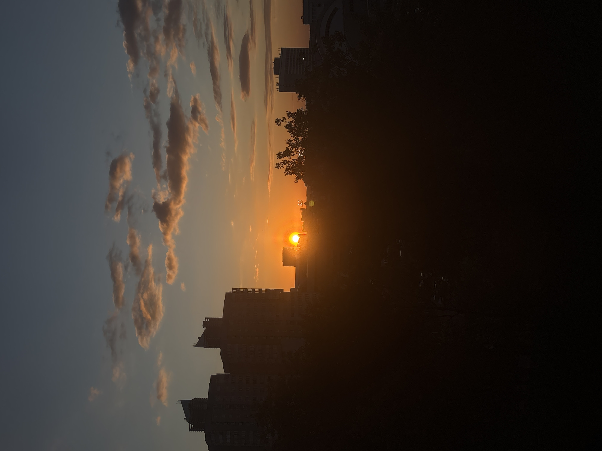 The image shows a sunset in Minsk, Belarus. The sun is low in the sky, casting a bright orange glow. The sky is a mix of blue and orange, with clouds tinged with pink and gold. Silhouetted buildings are visible in the foreground and middle ground. The right side of the image is predominantly dark, possibly due to trees or other structures blocking the light.