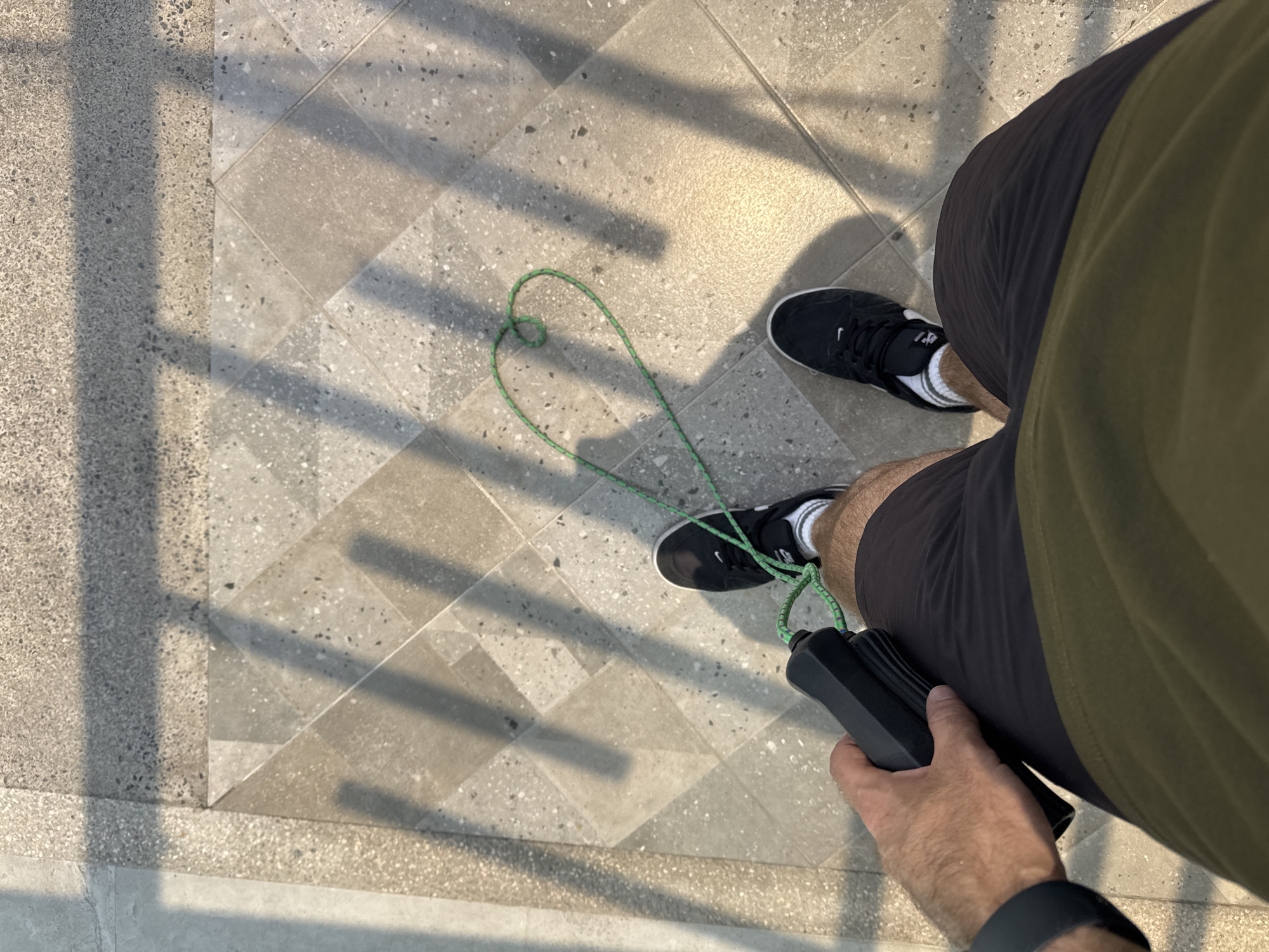 A top-down shot shows a person standing on a tiled floor with sunlight casting shadows. The person is wearing black athletic shoes with white socks, dark athletic shorts, and an olive-green shirt. They are holding a black jump rope with a green cord, which is arranged in a heart shape on the floor. A black smartwatch is visible on the person's wrist. The floor consists of square and rectangular tiles in varying shades of gray. This scene likely occurs in the city of San Bernardino Tlaxcalancingo, Mexico.