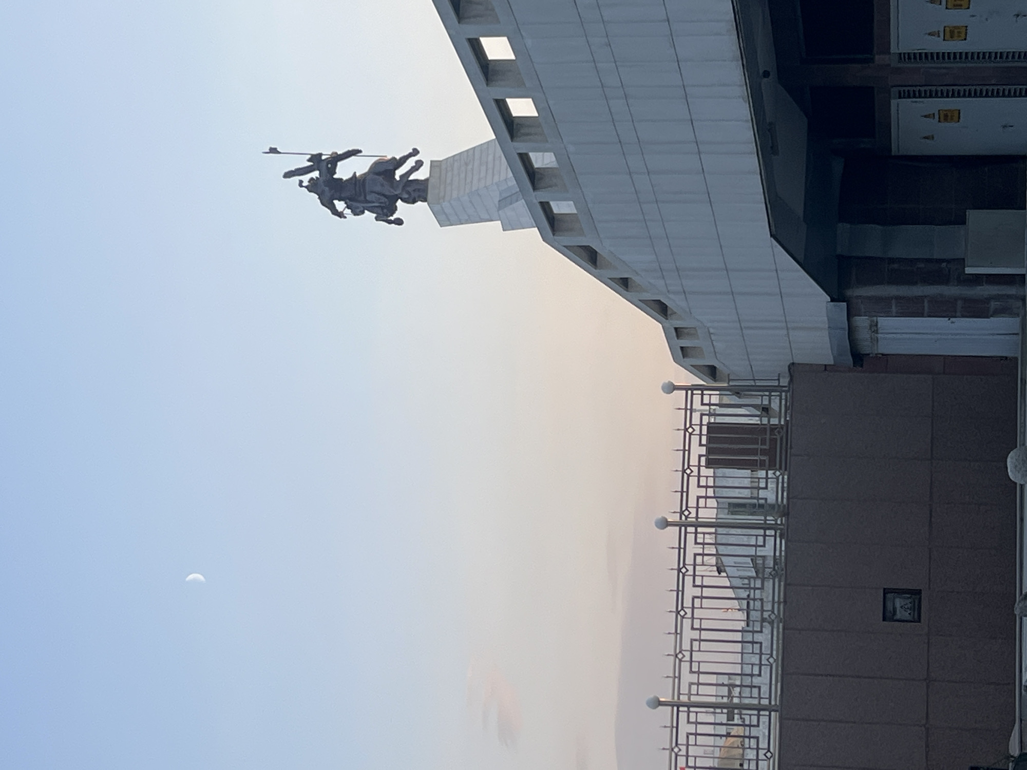 An outdoor, eye-level, medium shot captures the Victory Monument in Bishkek, Kyrgyzstan, under a pale blue sky with a sliver of a crescent moon. The monument, a dark bronze statue of a woman holding a wreath and a Soviet flag, stands atop a tiered pedestal. The foreground is dominated by a modern building with a white, angular facade and a dark-tiled lower section. A decorative metal railing runs along the edge of the foreground structure. The scene is devoid of any people or animals.