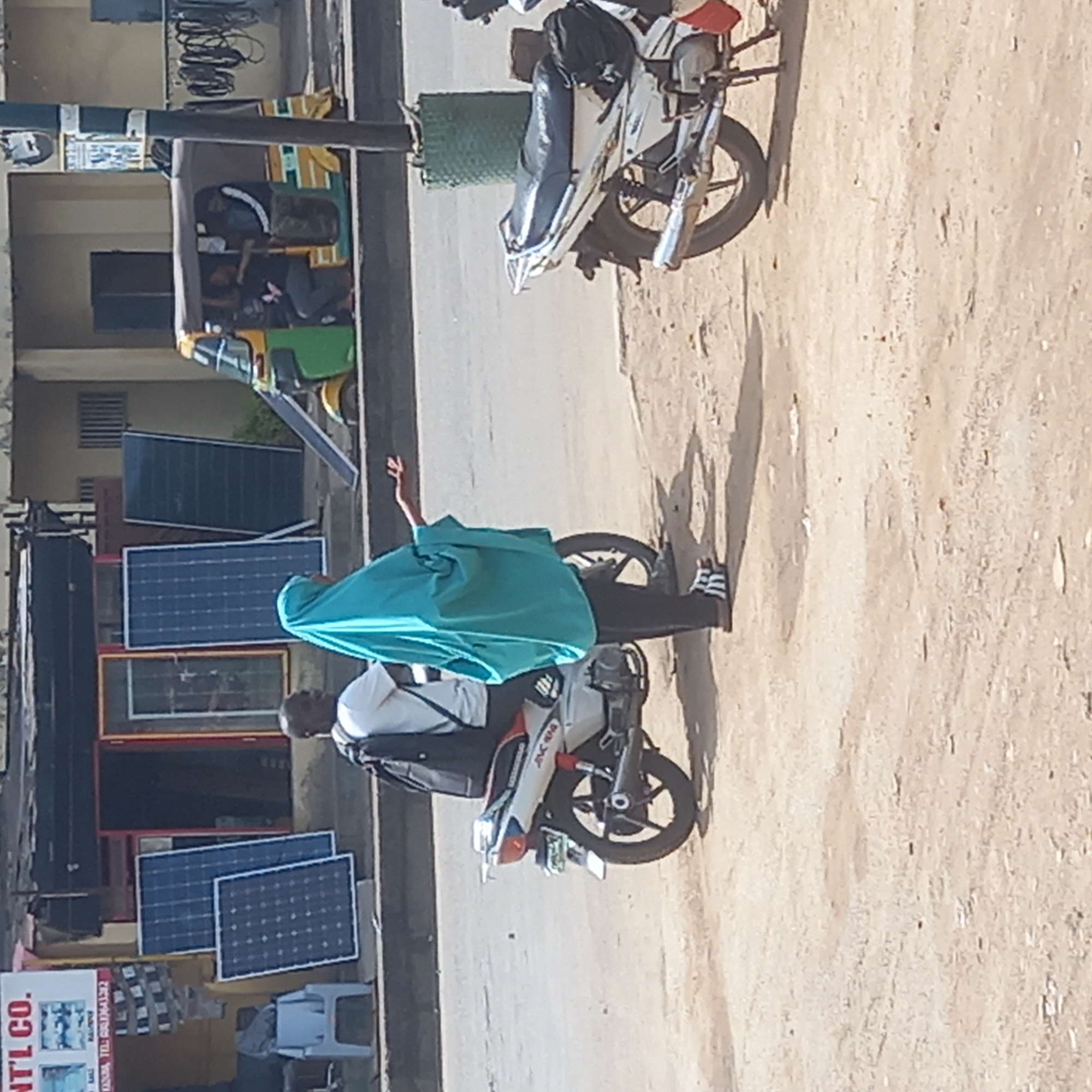 The image shows a street scene in Dadi Riba, Nigeria. A man is riding a motorcycle with a passenger wearing a blue head covering. The man is wearing a white shirt and black jacket. Solar panels are leaning against a building next to the motorcycle. A green and yellow auto rickshaw is parked nearby. Another motorcycle is parked on the side of the road. The ground is dusty.