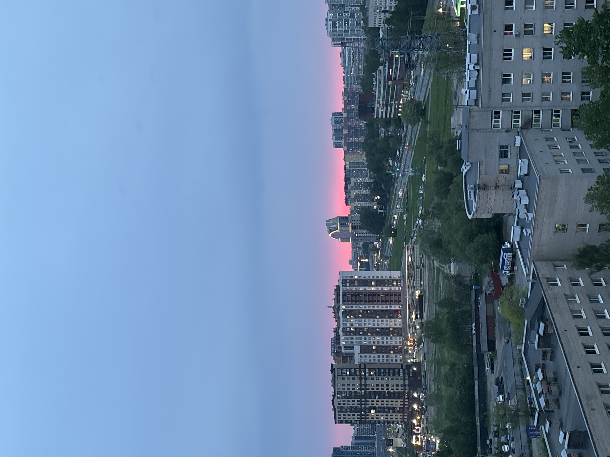The image shows a city at twilight. The sky is a gradient of blue and pink. Buildings of varying heights are visible, some with lit windows. A road runs through the city, with cars on it. There are trees along the road. The location is a city, but the specific city is unknown.
