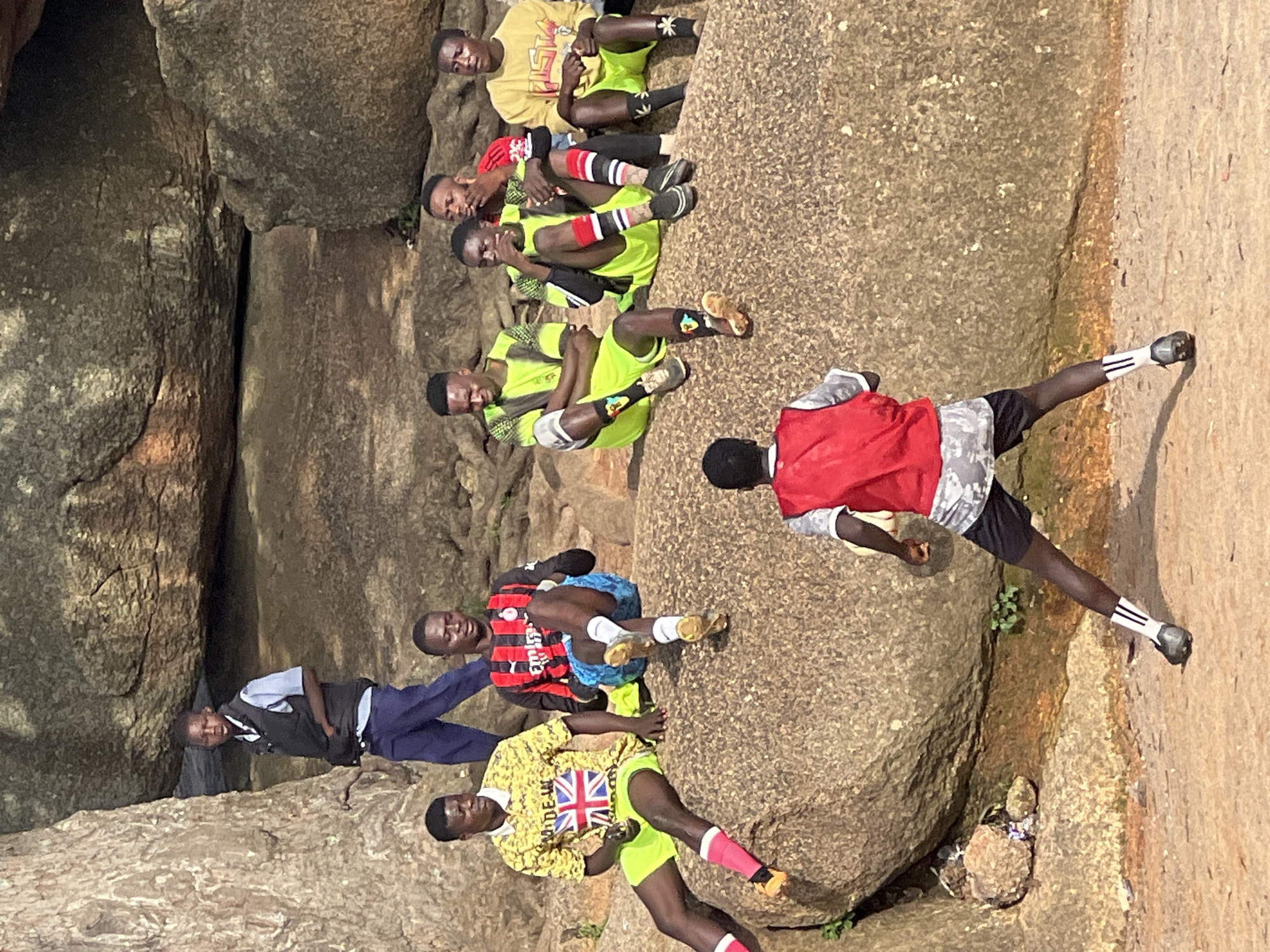 This image depicts a group of young men gathered on a rocky, outdoor terrain, possibly a hill or elevated area. In the foreground, one individual wearing a red vest and black shorts with white-striped socks is facing away from the camera, appearing to be in the process of kicking a soccer ball, though the ball is not visible. Behind him, seated on a large rock are seven other individuals, some in athletic wear. To the far left, a young person in a dark vest over a light blue shirt and dark blue pants stands by a large tree trunk. The environment suggests a casual gathering or a sports practice session in a natural, rocky landscape. The presence of soccer attire hints at a sporting context. The image appears to have been taken during daylight. It is impossible to determine the exact location within Jos, Nigeria, from the visual information provided.