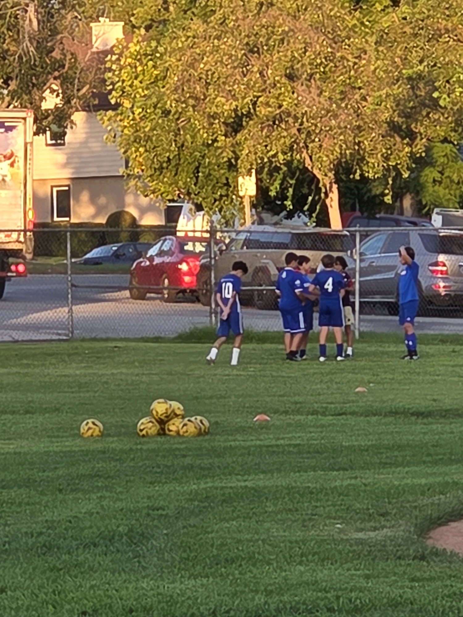 In Glendale, United States of America, a group of young boys in blue soccer uniforms are gathered on a grassy field, some with their arms around each other. One boy on the left wears a jersey with the number 10. Scattered on the grass are several yellow soccer balls. In the background, a chain-link fence separates the field from a street where cars are parked and driving. Behind the fence, houses and trees are visible, with one house featuring a white chimney. The lighting suggests it is late afternoon or early evening, with the sun casting a warm glow on the scene.