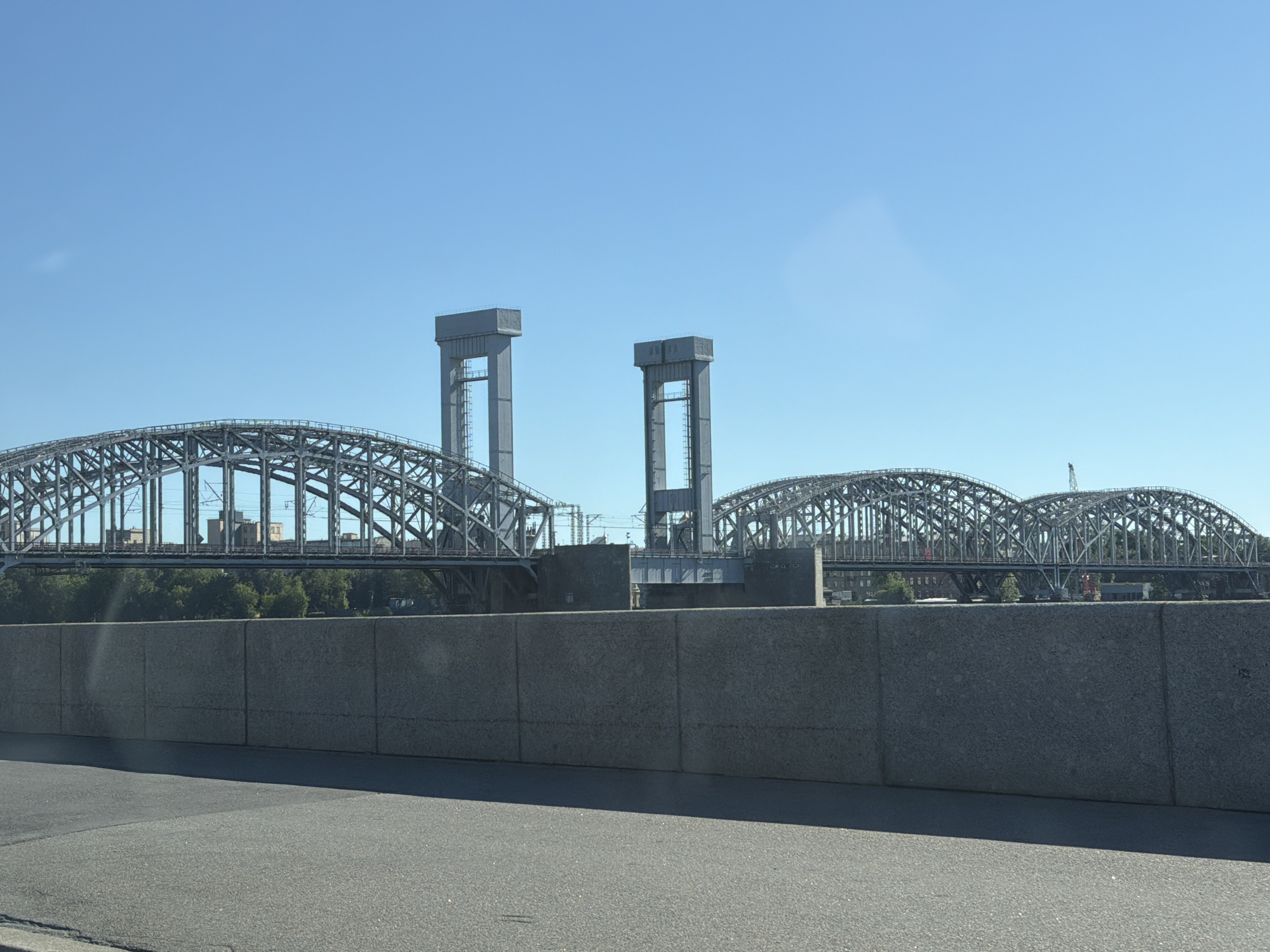 The image shows the Bolsheokhtinsky Bridge in Saint Petersburg, Russia, on a clear day. The bridge is a steel arch bridge with two large, distinctive vertical towers. The bridge spans across the Neva River. In the foreground, there is a grey concrete wall and a section of asphalt road. The background shows buildings and trees behind the bridge. The sky is a clear blue. There are no people or vehicles visible in the image.