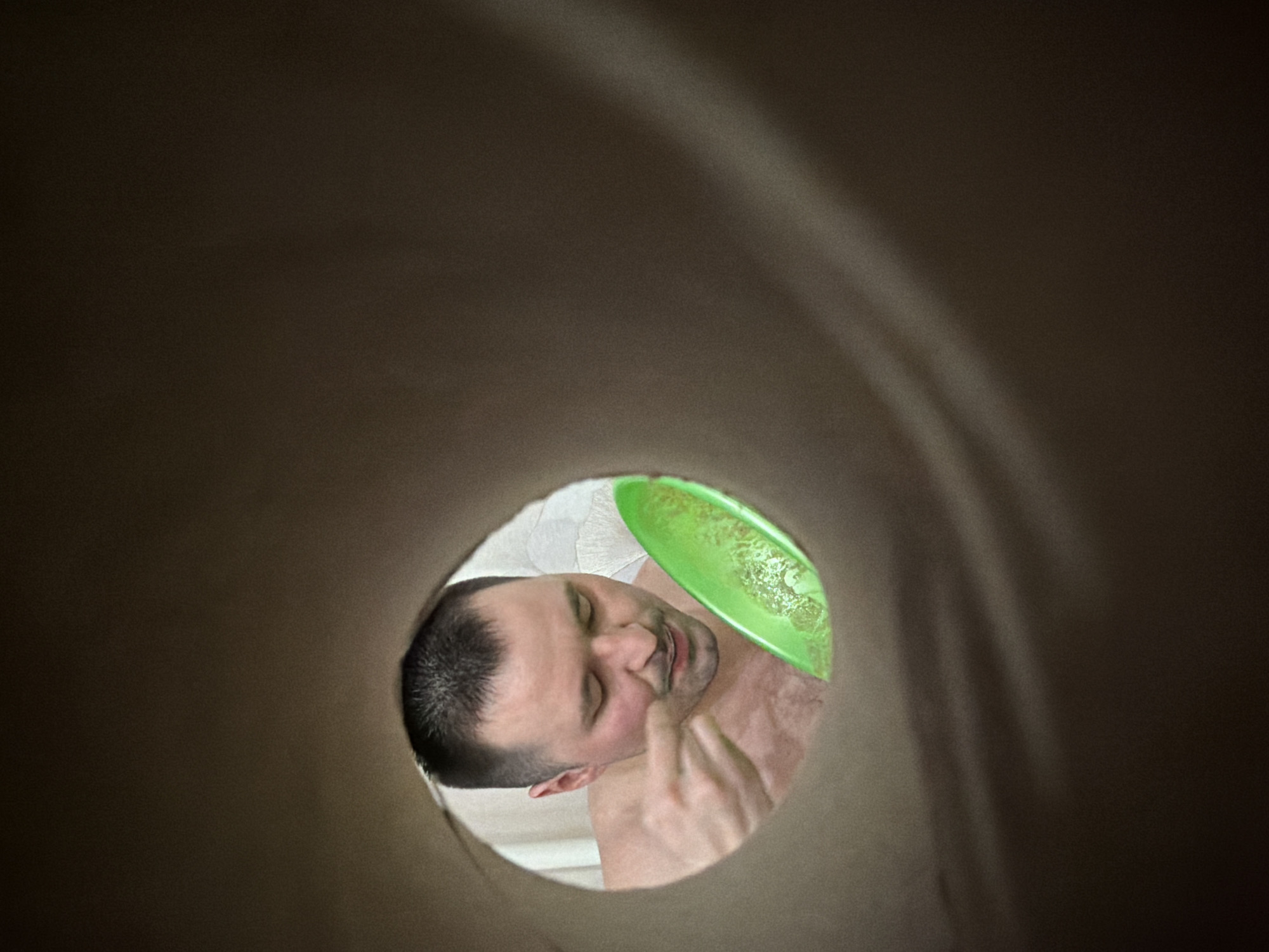 A man is seen through a circular opening inside a brown tube. The man has short dark hair, closed eyes, and is shirtless. He is holding a green plate or disc near his face. The background seen through the opening appears to be a white surface. The context of Izhevsk, Russia is not evident from the provided image.