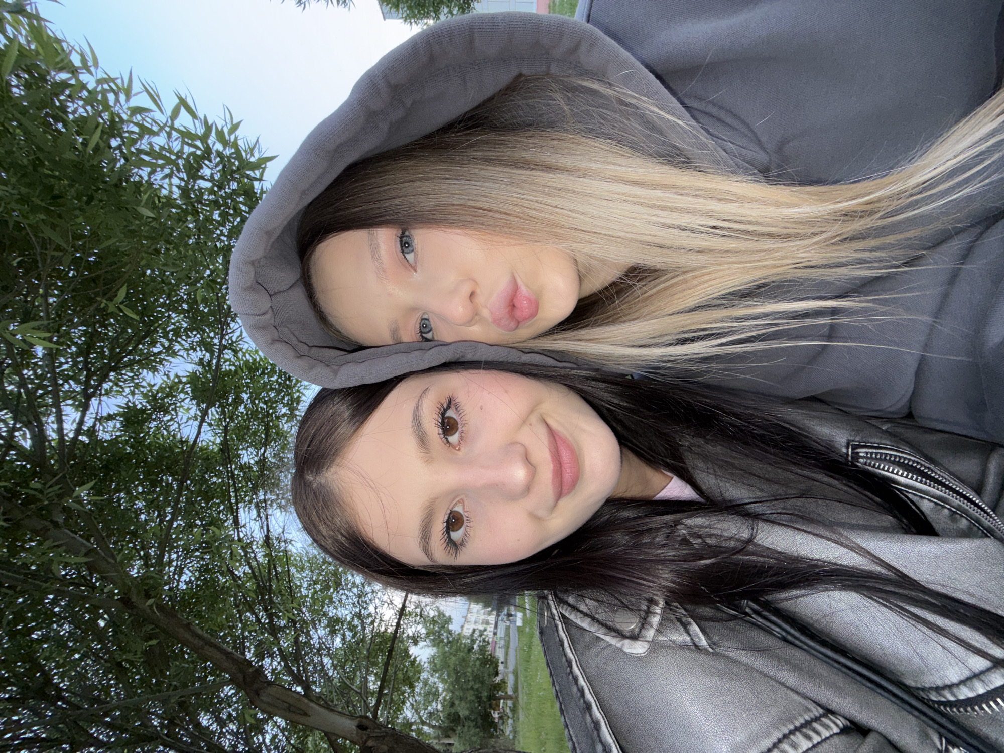 Two young women are posed closely together for a selfie outdoors. The first woman has fair skin, blonde hair, and blue eyes; she is wearing a gray hoodie and puckering her lips. The second woman has fair skin, dark hair, and brown eyes; she is smiling and wearing a gray leather jacket. The background shows trees and a building, possibly in Chaikovsky, Russia.