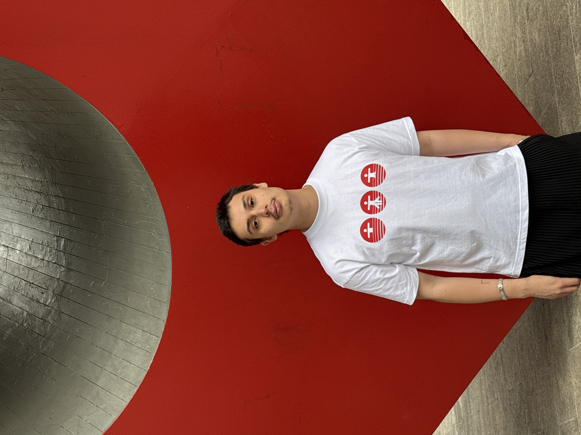 The image captures a man wearing a white t-shirt with three red emblems. He is standing in front of a curved, silver-colored structure set against a red wall. The background suggests an interior architectural element, possibly part of a public space, with an adjacent grey surface on the right. The location, based on the architectural style, is likely in the city of New York, United States of America.