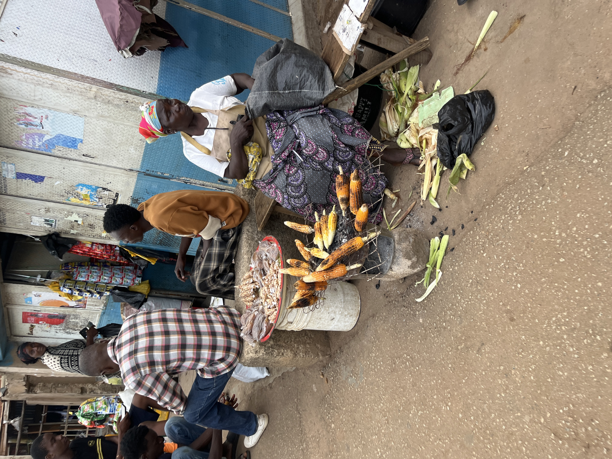 In the city of Bukuru, Nigeria, a street scene unfolds. A woman wearing a colorful headscarf, a white t-shirt, and a patterned skirt sits on a wooden bench. She is engaged in preparing or selling food, with a collection of corn cobs on a metal grill in front of her. Next to her is a large black plastic bag and a table with some paper items. Behind her, more corn husks and what appear to be bunches of scallions are scattered on the ground. In the foreground, several individuals are seated or crouching, some appearing to be customers. One man in a plaid shirt is sitting on a concrete block next to a red platter filled with small, packaged food items and what looks like dried seafood. The background shows a building with a blue and grey facade, adorned with posters and what might be a shopfront. The ground is a dirt road.