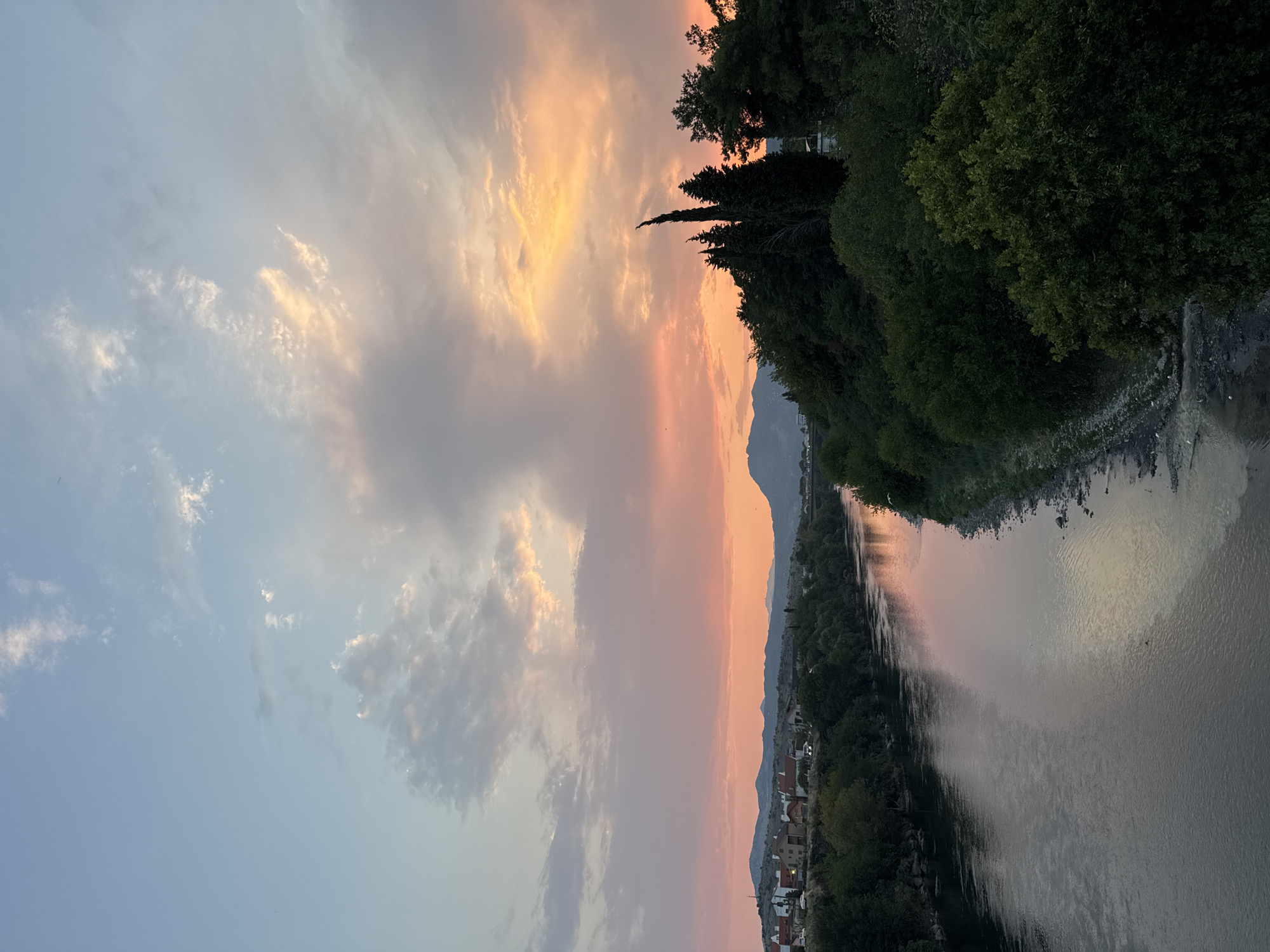 An aerial, eye-level view captures a wide, calm river flowing through a verdant landscape at sunset. The sky is a canvas of soft pinks, oranges, and blues, with wispy clouds catching the fading light. On the left bank, a line of buildings with red-tiled roofs forms a small town, nestled against rolling hills. The right bank is densely covered with lush green trees, including a few tall, dark cypress trees reaching towards the sky. The river reflects the colors of the sunset, creating a shimmering pathway of light. The water appears still, with only slight ripples disturbing its surface. The scene conveys a sense of tranquility and natural beauty as day transitions into night.