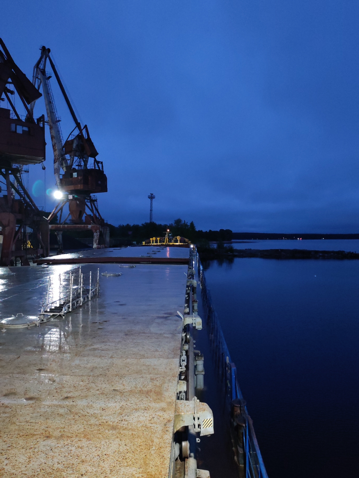 The image presents an industrial port scene at dusk or night, viewed from the deck of a vessel, likely a cargo ship or barge, in Medvezhyegorsk, Russia. The ship's deck in the foreground is wet and appears worn with rust, reflecting artificial lights.
Two large, rusty port cranes dominate the left side of the mid-ground. One crane is brightly illuminated by its own light, and the number "43" is visible on its structure. Reflections from these lights are prominent on the wet deck. Beyond the cranes, a tall, slender tower stands against the dark blue sky, and other smaller, yellow industrial equipment can be seen on the distant dock.
To the right, a dark body of water stretches towards a faint shoreline in the distance, where a few small lights are discernible. The sky above is a deep, rich blue, characteristic of twilight or nighttime conditions. No people are visible in the frame, contributing to a quiet, stark, and industrial atmosphere. Yellow and black striped hazard markings are present on machinery near the ship's railings. The wet surfaces suggest recent rain or a damp environment.