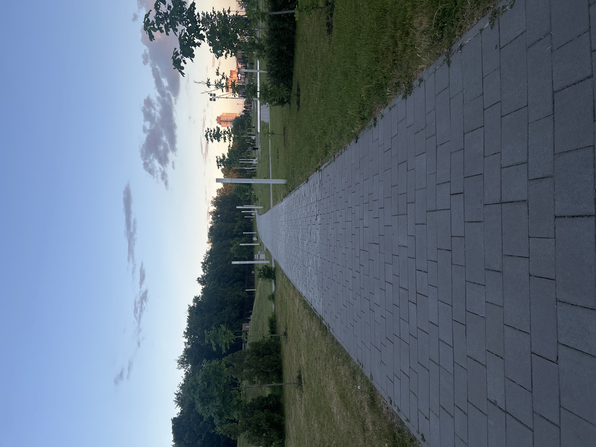 The image shows a paved walkway in a park setting. The walkway, constructed of gray rectangular pavers, leads into the distance. To the left of the walkway is a strip of green space with trees and bushes. To the right is a grassy area. In the background, buildings are visible. The sky is blue with some clouds. The location is likely Tolyatti, Russia. White posts are lined on the left side of the walkway. The time of day appears to be late afternoon or early evening. No people are visible in the image.