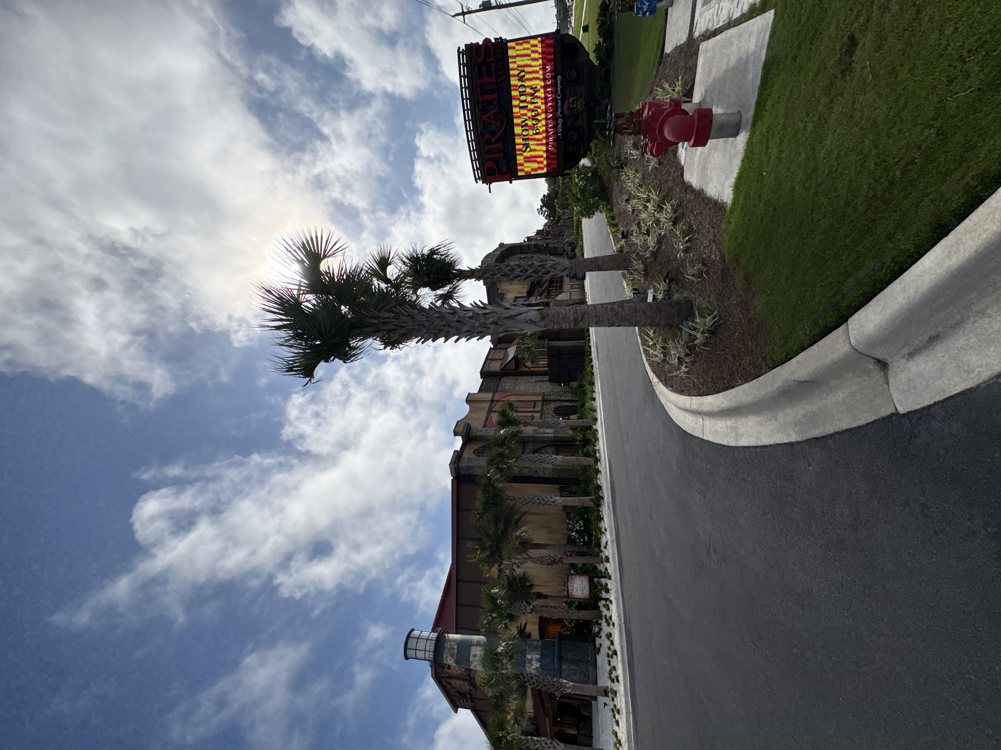 The image depicts Pirates Voyage Dinner & Show in Panama City Beach, Florida, United States. The view is from street level, angled upwards to showcase the building's facade and a large marquee sign advertising the show. A palm tree is positioned in the foreground, partially obscuring the sun. A red fire hydrant stands near a grassy area and a concrete walkway. The sky is blue with scattered clouds. A paved road occupies the lower right portion of the frame.