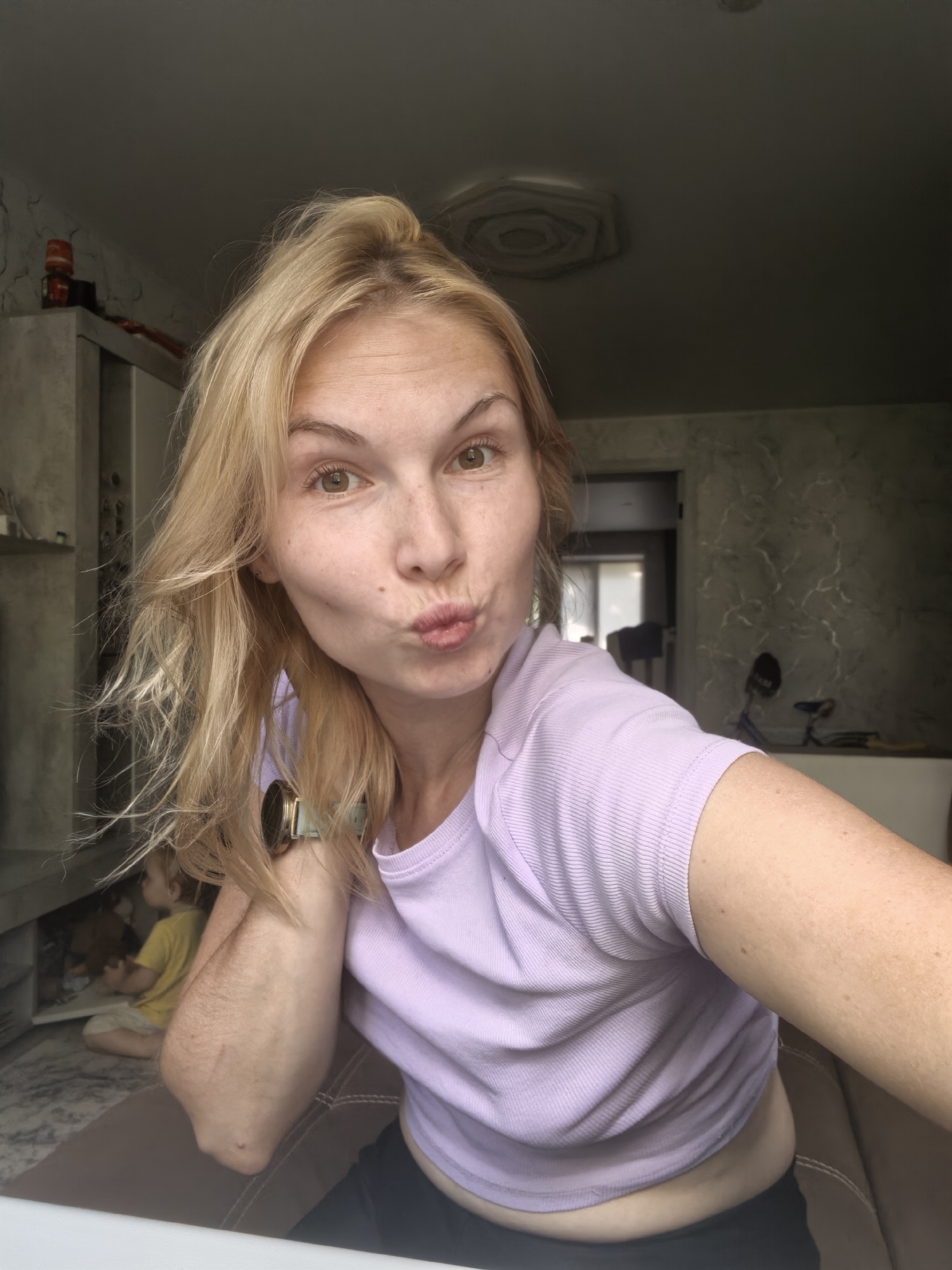 A blonde woman with light brown eyes and freckles on her cheeks and nose is pouting her lips in a selfie. She has her hair pulled up in a messy bun with some strands falling around her face. She is wearing a lavender t-shirt and a watch on her left wrist. The background shows a room with furniture, including a cabinet and a sofa, and a young child in a yellow shirt sitting on the floor. The image appears to be taken indoors, possibly in a residential setting in Sterlitamak, Russia.