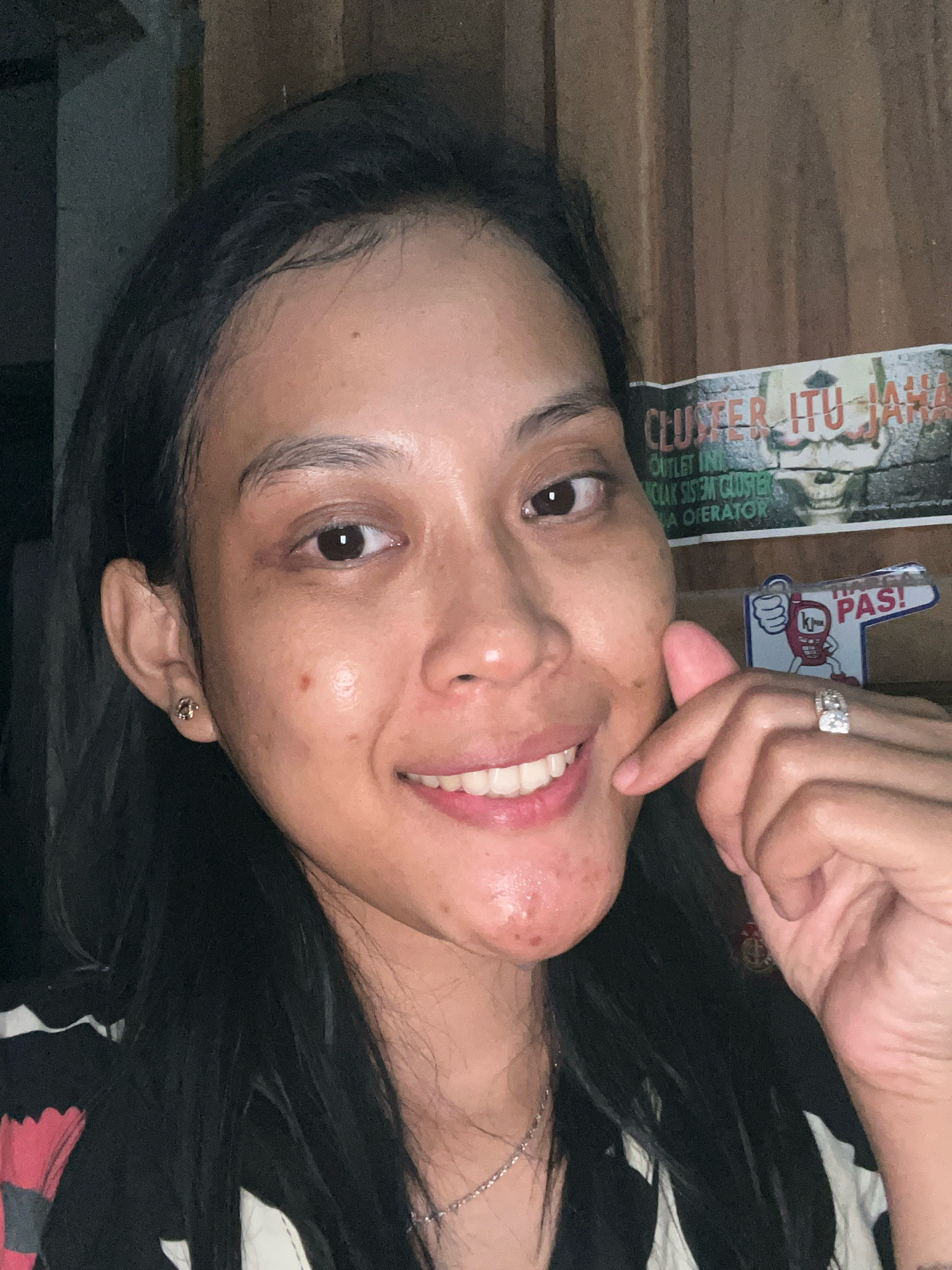 A young woman with dark hair and fair skin is taking a selfie indoors. She is smiling, and her face has some visible acne. She is wearing a black and white striped shirt and a necklace. On her left ear, she has a small stud earring. On her right hand, she is wearing a ring. In the background, there is a wooden wall and a poster with text in Indonesian, which mentions "CLUSTER ITU JALAN". To the right of the woman, there is another small sign that says "HARGA PAS!". The overall lighting suggests it is nighttime or a dimly lit room. The location is specified as Kecamatan Pati, Indonesia.
