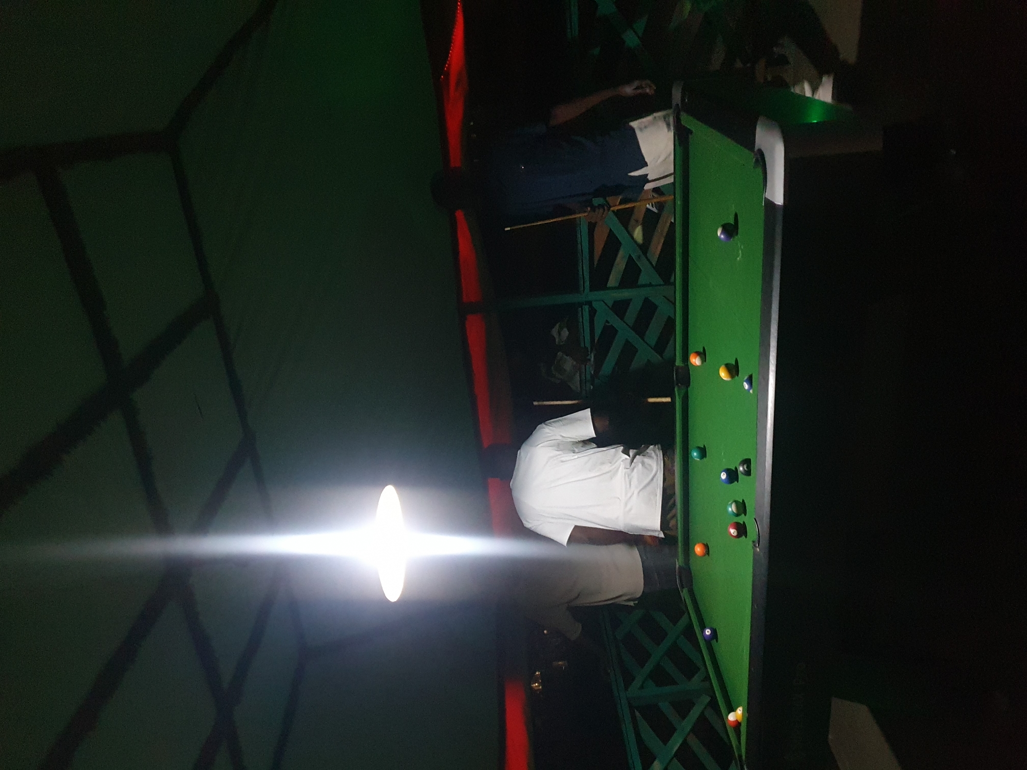The image shows two people playing pool in a dimly lit setting, possibly at night. One person, wearing a white t-shirt, is standing near a pool table with several balls on it. Another person, wearing dark blue pants and a white shirt, is also visible near the table, holding a pool cue. The pool table is green and appears to be outdoors, with a green lattice structure around it. There's a bright light source in the background, casting a glare across the scene. The overall atmosphere is dark, suggesting it's nighttime. There are no visible signs or text in the image. The context suggests this is Lokoja, Nigeria.