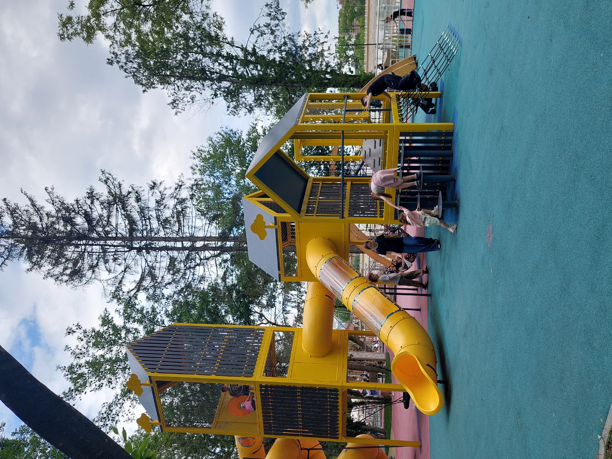 An aerial view of a bright yellow playground with a tubular slide, climbing structures, and a netted enclosure. Several children are playing on the equipment, some climbing, others sliding. The playground is set on a blue and red soft-surface ground, surrounded by trees. In the background, a body of water and a cityscape are visible, hinting at the location in Alikonovka, Russia. The sky is overcast, with scattered clouds.
