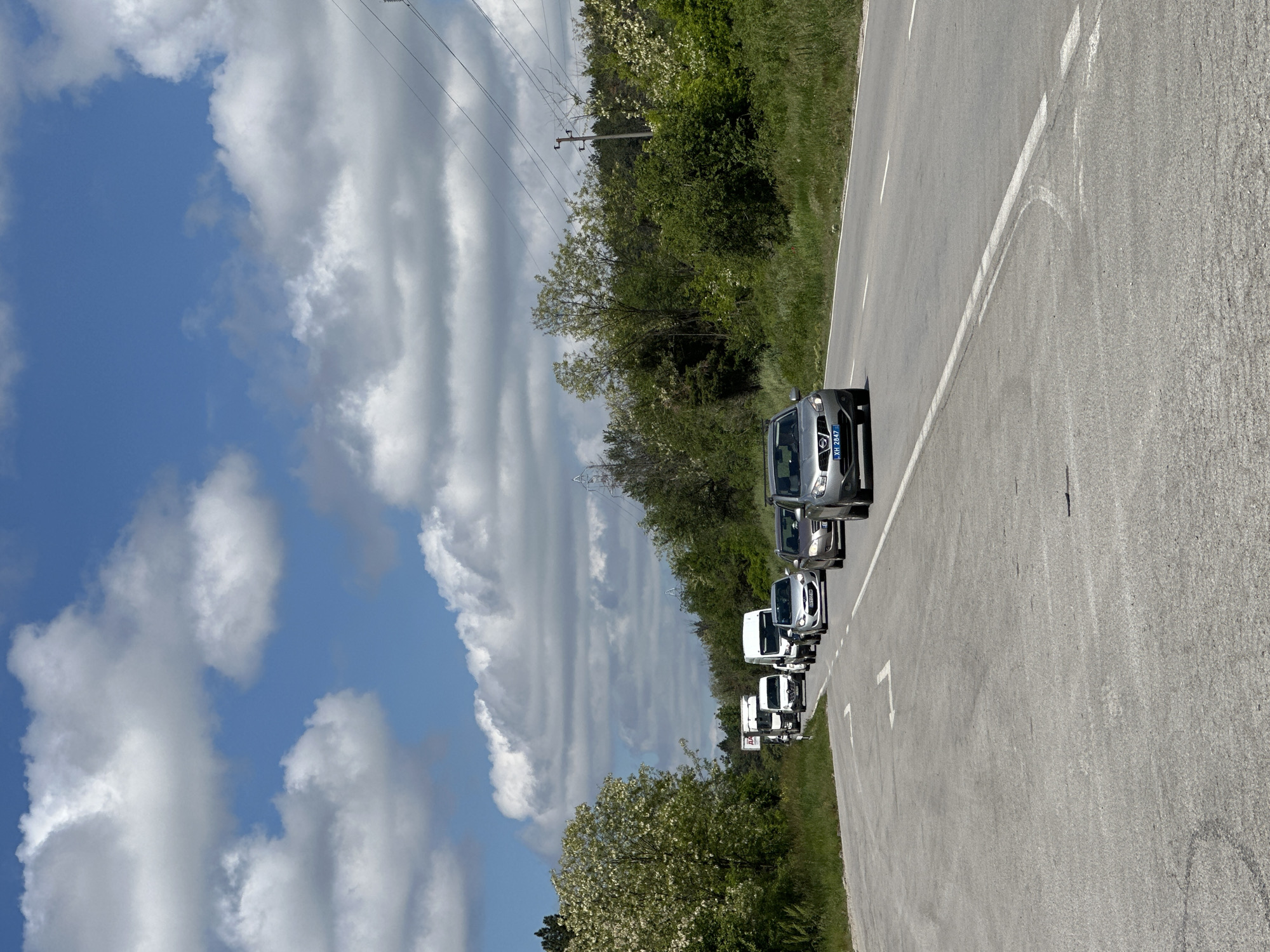 The image depicts a convoy of vehicles traveling on a road, potentially near the city of Ledenik, Bulgaria. There are several cars visible in a row, indicating a planned trip or parade. The environment includes trees and greenery along the roadside, with power lines present. The sky is filled with clouds. The road surface is asphalt, marked with white lines. No people are explicitly visible.