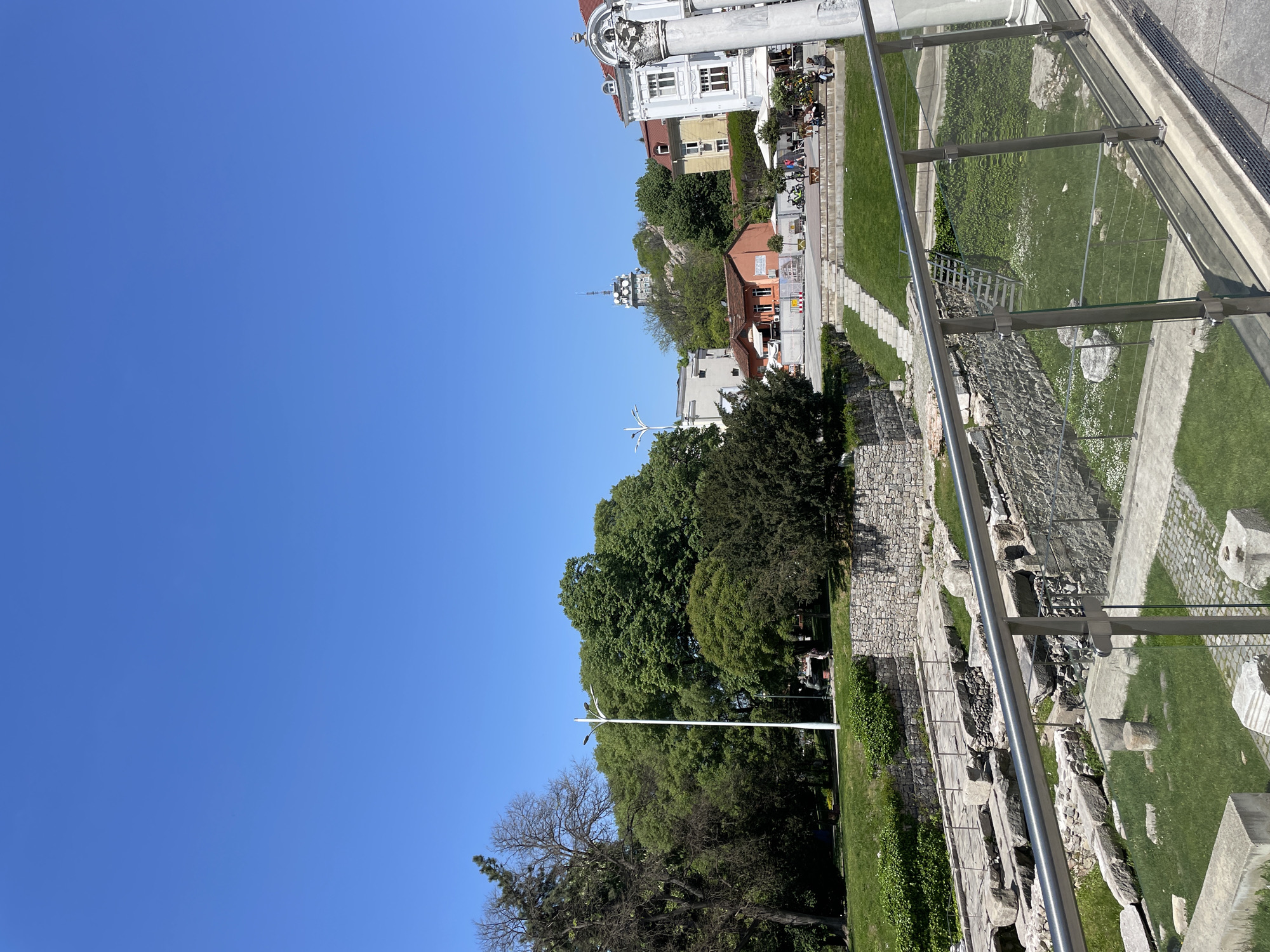 The image, taken in Plovdiv, Bulgaria, depicts a panoramic view of a historical city with modern elements, under a clear blue sky. The camera appears to be looking down from an elevated position, possibly from a viewing platform or higher ground, with a glass and metal railing running diagonally across the foreground, suggesting a public viewpoint.
In the immediate foreground and mid-ground, there are visible stone foundations and walls, characteristic of ancient ruins or an archaeological site, interspersed with grassy areas. These ruins suggest a rich historical past for the location. A set of stone steps leads down into the archaeological area.
Beyond the ruins and green spaces, a vibrant urban landscape unfolds. Numerous lush green trees, some tall and mature, provide ample shade. A tall, white, multi-armed lamppost stands prominently amidst the trees. Further in the mid-ground, several buildings are visible: a large, ornate white building with a red roof and classical architectural details (columns, statues) stands out towards the upper right. Nearby, a smaller, rustic-looking building with an orange-red roof is nestled among other structures. On a terrace to the right of the white building, several people are seated at outdoor tables, enjoying the sunny weather, indicative of a cafe or restaurant.
In the distant background, atop a hill, a distinctive modern communication tower or antenna structure can be seen, providing a contrast to the historical elements below. The sky is a uniform, brilliant blue, completely clear of clouds, indicating a bright and sunny day, likely mid-morning or early afternoon given the strong lighting and shadows. No discernible text is visible in the image.