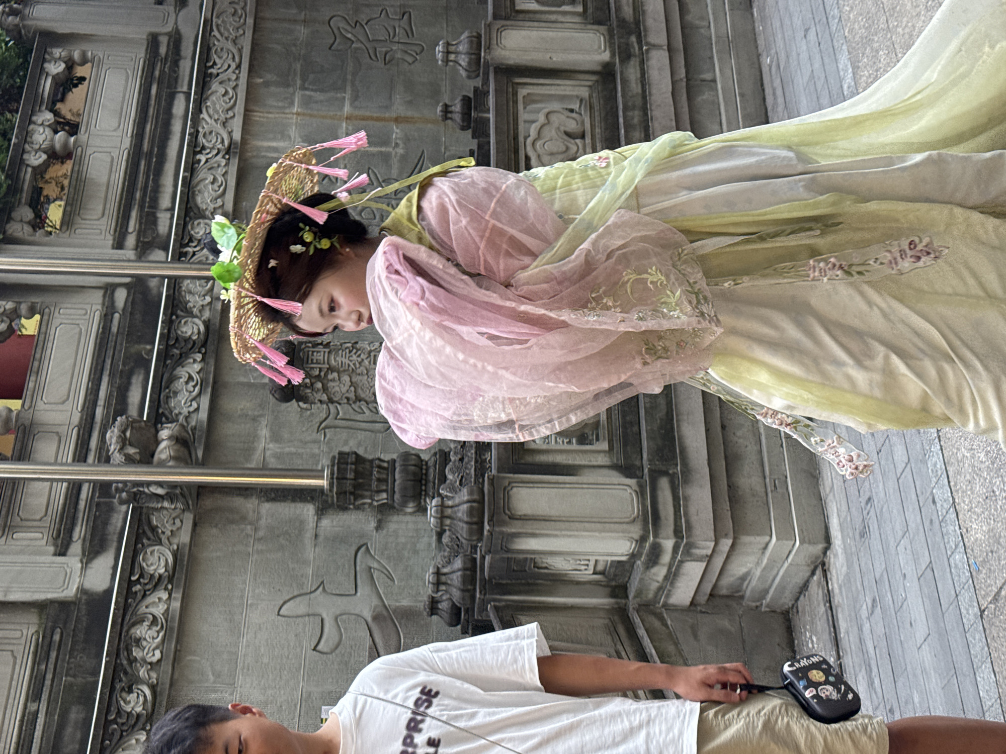 In the Yuzhong District of China, a young East Asian woman adorned in traditional Hanfu, featuring pale green and pink layered garments and a straw hat with pink tassels, walks past an East Asian male wearing a white t-shirt and beige shorts, who is holding a small black pouch. The backdrop consists of intricate, weathered stone architecture with carved motifs.