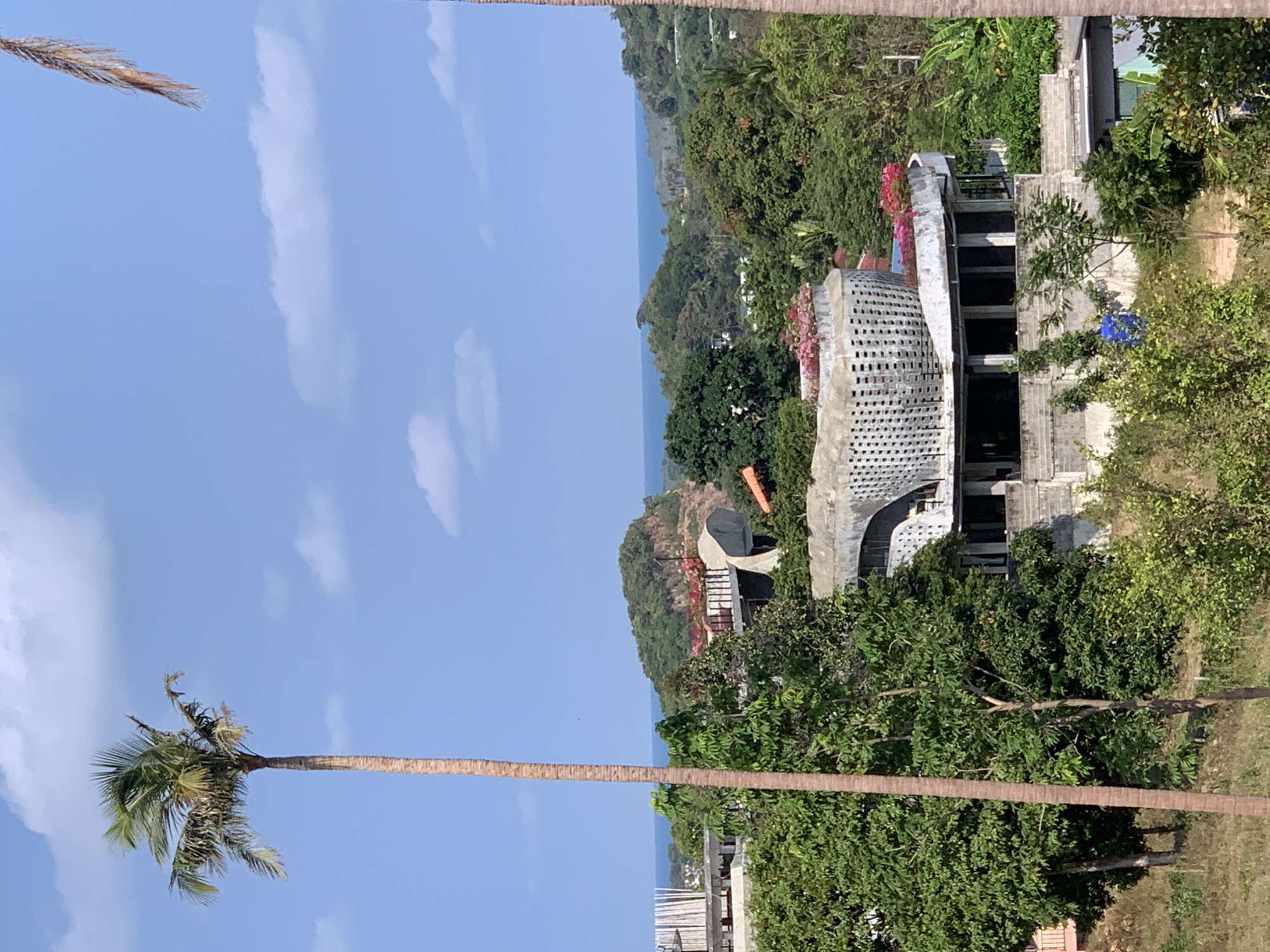 The image shows a coastal scene near บ้านบางรักษ์, Thailand. In the foreground, a tall palm tree stretches across the frame against a blue sky with scattered clouds. The middle ground features a building with a distinctive modern architectural design, characterized by a white perforated exterior and curved structure, nestled among dense green trees and vegetation. The background reveals a glimpse of the sea, visible between the trees, and the distant shoreline.