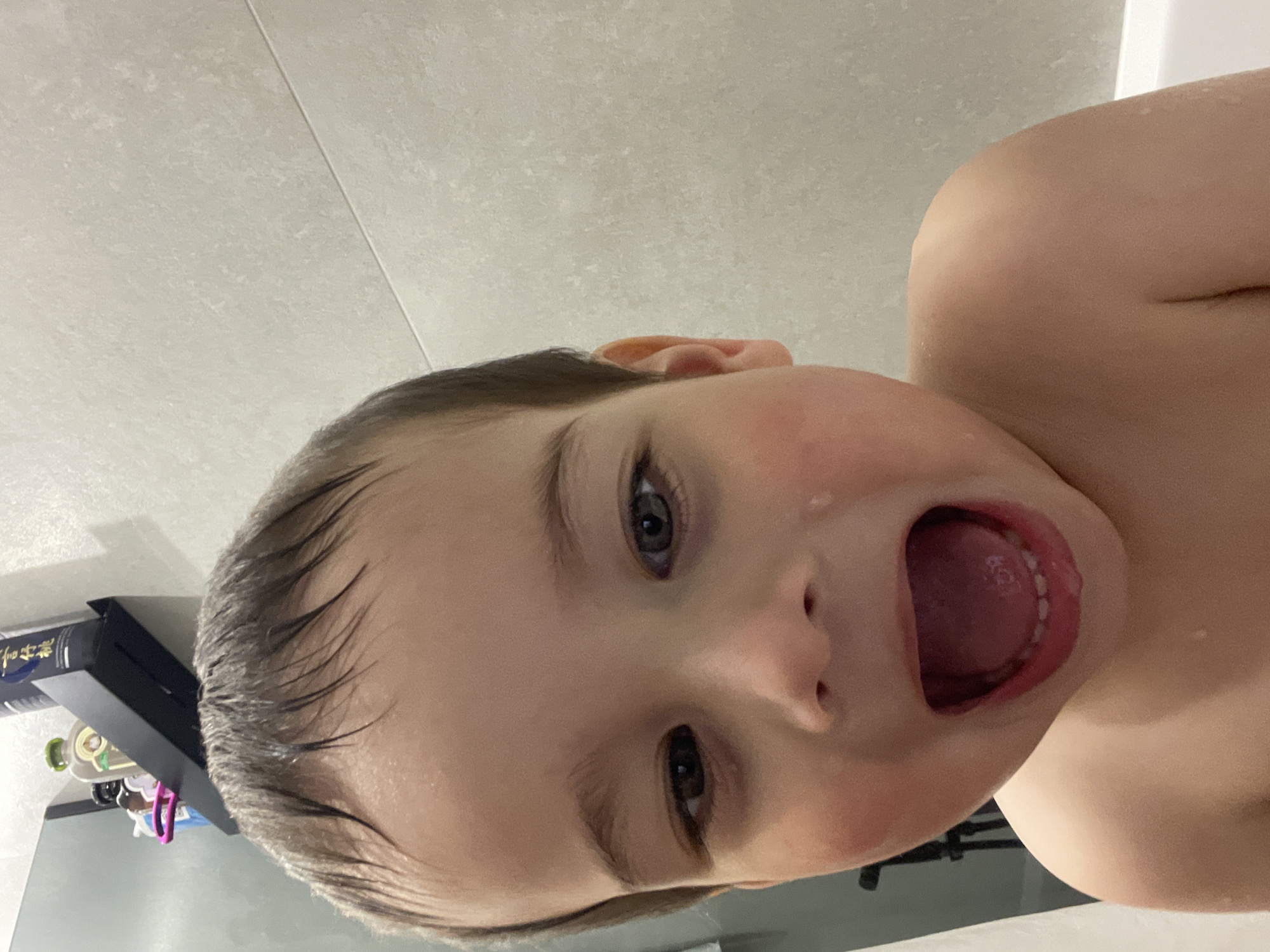 A young child with wet hair is looking upwards and opening their mouth wide, possibly shouting or reacting to something unseen. The child appears to be in a bathroom setting, with light-colored tiles visible in the background and a shelf or ledge with various bottles and items to the side. It is not possible to determine the location of Izhevsk, Russia, or the presence of subjects and interactions from the image.