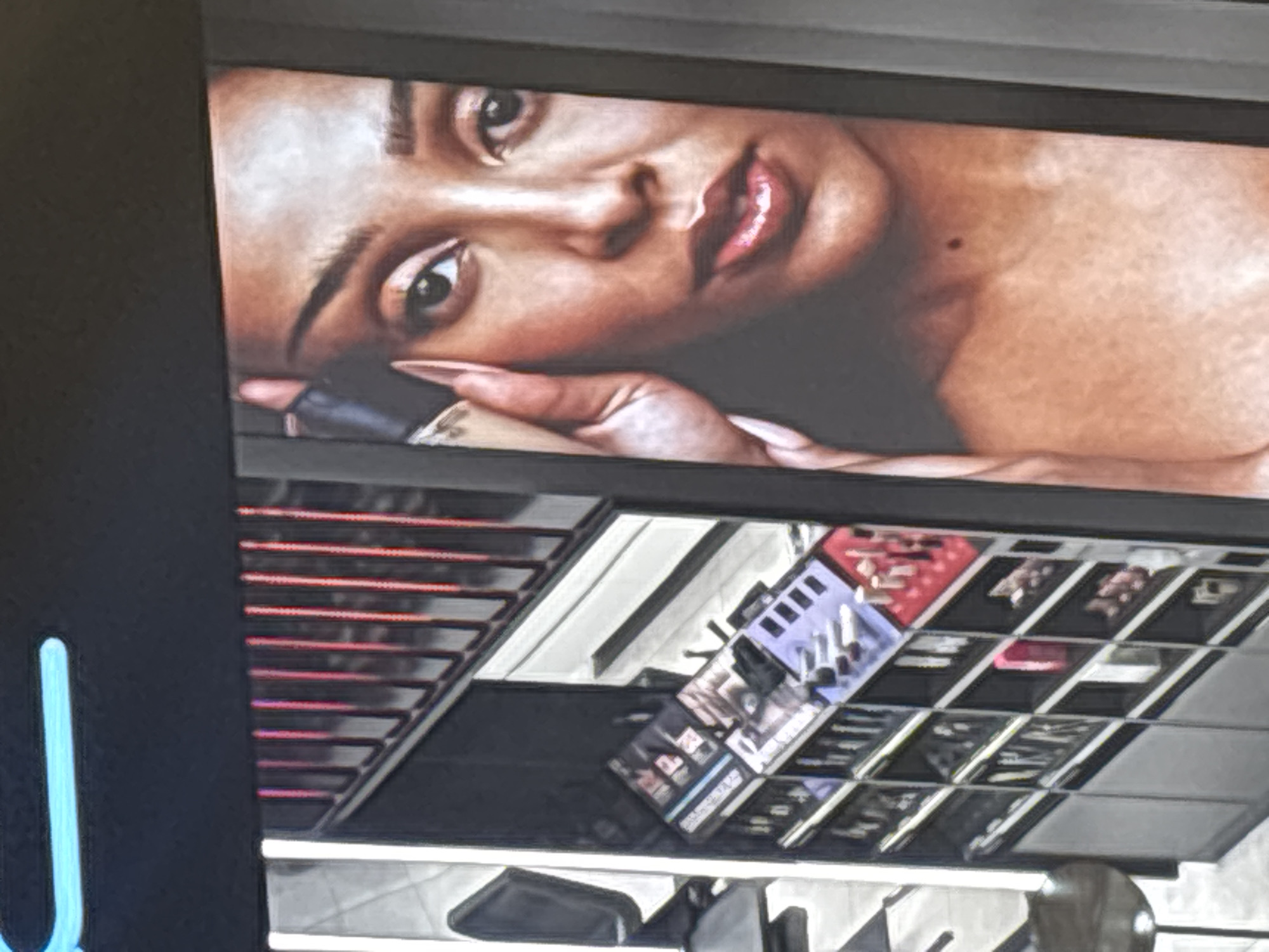 The image shows a makeup display in what appears to be a retail setting. Above the display is a large advertisement featuring a woman with brown skin lying on her side, holding a makeup bottle, likely promoting foundation. Below the advertisement, there is a makeup display with various products, arranged in a grid pattern with different colors and types of makeup visible. Chairs and possibly mannequin heads are visible beneath the display, suggesting it is located within a store. The exact location in Yerevan, Armenia, cannot be determined from the image alone.