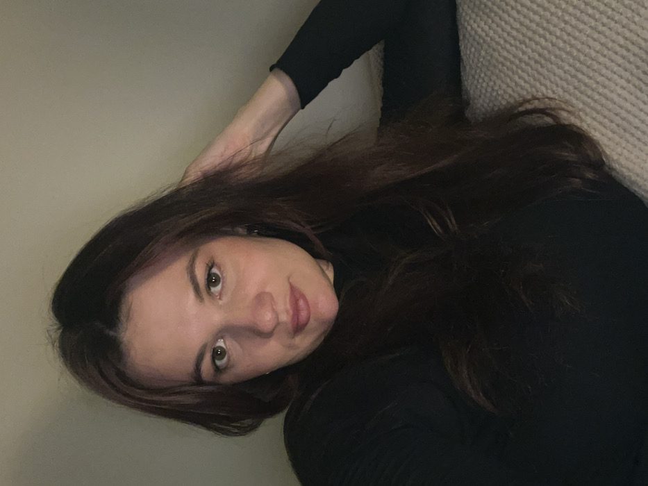 A woman with long, dark brown hair is lying on a light-colored couch. She wears a black, long-sleeved turtleneck shirt. One arm is bent behind her head, resting on a light beige wall. She gazes directly at the camera with a slight smile. The lighting is somewhat dim.