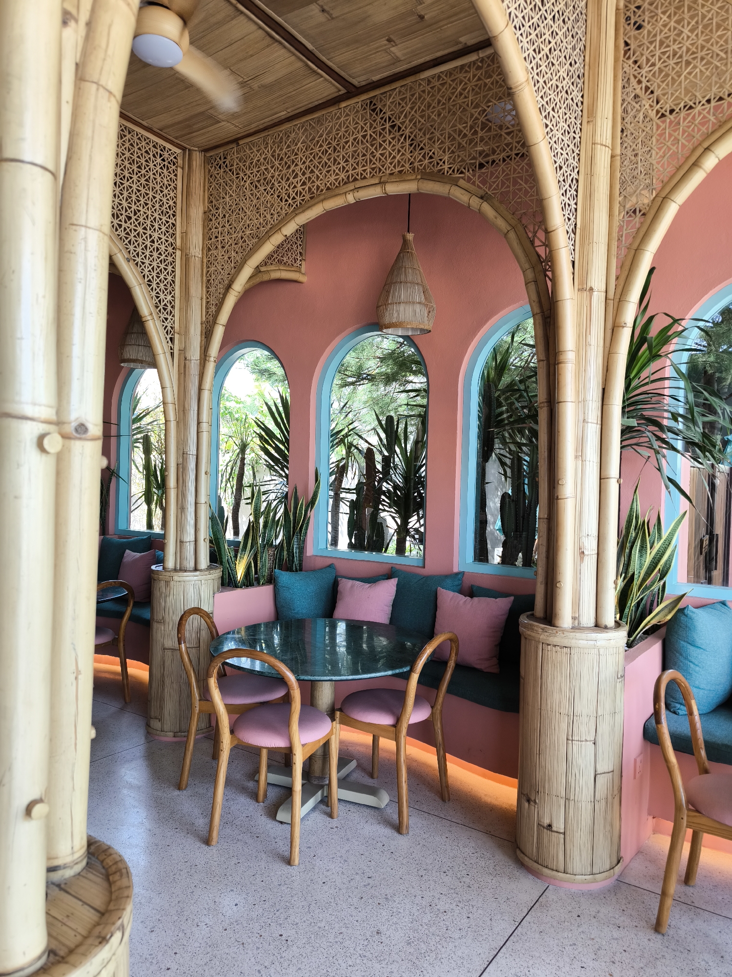 The image shows the interior of a stylish cafe or restaurant in Unknown, Indonesia. The design features a color palette of salmon pink, turquoise, and natural bamboo. Three arched openings, framed in bamboo and pink walls with turquoise trim, reveal exterior views of greenery, including trees and cacti. The interior arches are decorated with woven bamboo panels.
Below the arched openings, cushioned booths are arranged, upholstered in teal and adorned with pink throw pillows. Round tables with dark green surfaces are surrounded by wooden chairs with pink cushions.
Bamboo is a dominant element in the design. The room is supported by bamboo columns. A woven bamboo lampshade hangs from the ceiling. The floor is speckled with shades of pink.
Several plants, including snake plants and palms, are arranged within the interior space, adding to the tropical vibe. The cafe appears to be empty of people.