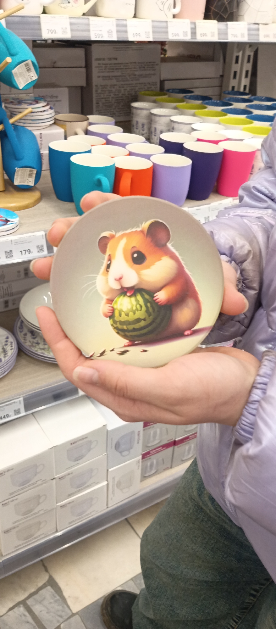 A person's hands are centered in the frame, holding a round, light-colored plate or coaster. The object features a charming illustration of a plump, brown and white hamster with large dark eyes, happily munching on a green watermelon. The person is wearing a light purple puffy jacket, visible on their right arm and shoulder, and blue jeans on their left leg.
The scene is an indoor retail environment, likely a homeware or kitchenware section of a store. Shelves are laden with various ceramic items. In the background, numerous colorful mugs in shades of blue, orange, purple, and pink are arranged. Other white and patterned cups are also visible, along with stacks of plates featuring a blue floral design. A wooden display stand with blue items hangs to the left.
Price tags are visible throughout the store display, showing numbers such as "179.", "799.", "599.", and "449.", some with accompanying QR codes. On a large cardboard box in the mid-background, Cyrillic text is faintly visible, with "Условия хранения" (Storage conditions) being partially legible. Smaller white boxes on the bottom shelf also show text, possibly brand names like "Kupi Luxe", and outlines of cups. The floor is tiled with light and dark grey squares. The activity suggests someone browsing or examining merchandise in a store.