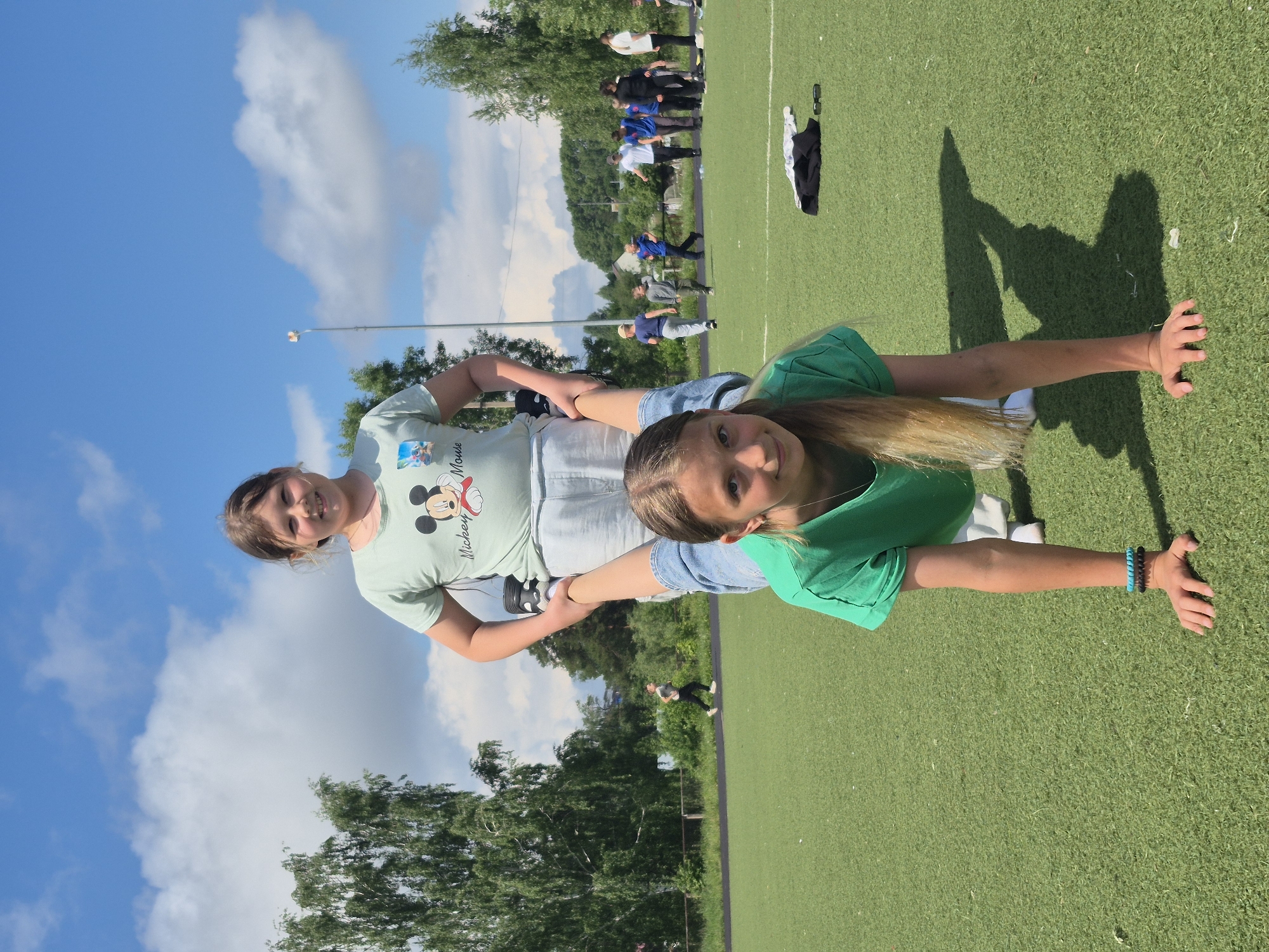 This image captures an outdoor scene on a bright, sunny day, likely at a sports field in Chaikovsky, Russia. In the foreground, two young girls are engaged in an acrobatic or balancing pose on artificial green turf. One girl, with long blonde hair and wearing a green t-shirt and blue denim shorts, lies on her back with her arms outstretched to the sides, supporting the other girl who stands on her bent legs/waist. This second girl wears a light-colored t-shirt featuring a "Mickey Mouse" graphic and light-colored shorts.
The girl in the green t-shirt smiles directly at the viewer, while the other girl looks towards the camera with a somewhat focused expression. Strong, distinct shadows are cast on the green field, indicating direct sunlight and suggesting it is likely mid-day or early afternoon. The sky above is a clear blue with scattered white clouds.
In the background, several other individuals are visible standing and walking on the sports field and an adjacent track. The field is bordered by numerous lush green trees under the clear sky. A street light pole is also discernible in the mid-ground. On the ground near the girl in the green shirt, a pair of sunglasses and some folded clothing are visible. The girl in the green shirt wears two beaded bracelets, one blue and one black, on her left wrist.