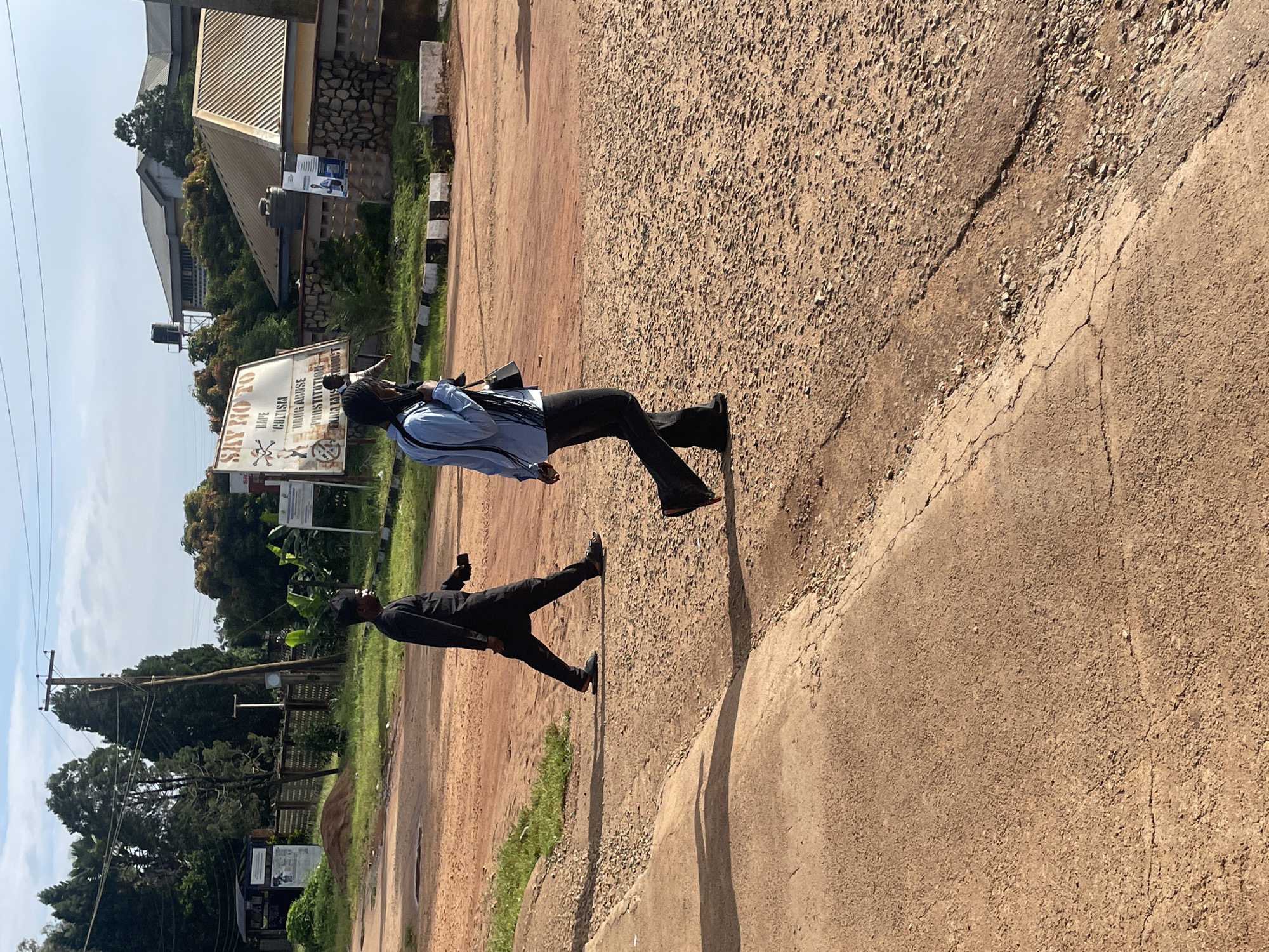 Two people are walking across a dirt and gravel road in front of some buildings. The person on the left is a man wearing a black robe and a black cap. He is walking away from the camera and holding a small object in his right hand. The person on the right is a woman wearing a light blue jacket, black pants, and a black headscarf. She is also walking away from the camera, and she has a dark bag slung over her shoulder. A sign with text and graphics is visible in the background, and there are trees and power lines in the distance. The environment appears to be a somewhat undeveloped area, possibly on the outskirts of a city. The road surface is uneven with patches of dry grass and small stones.