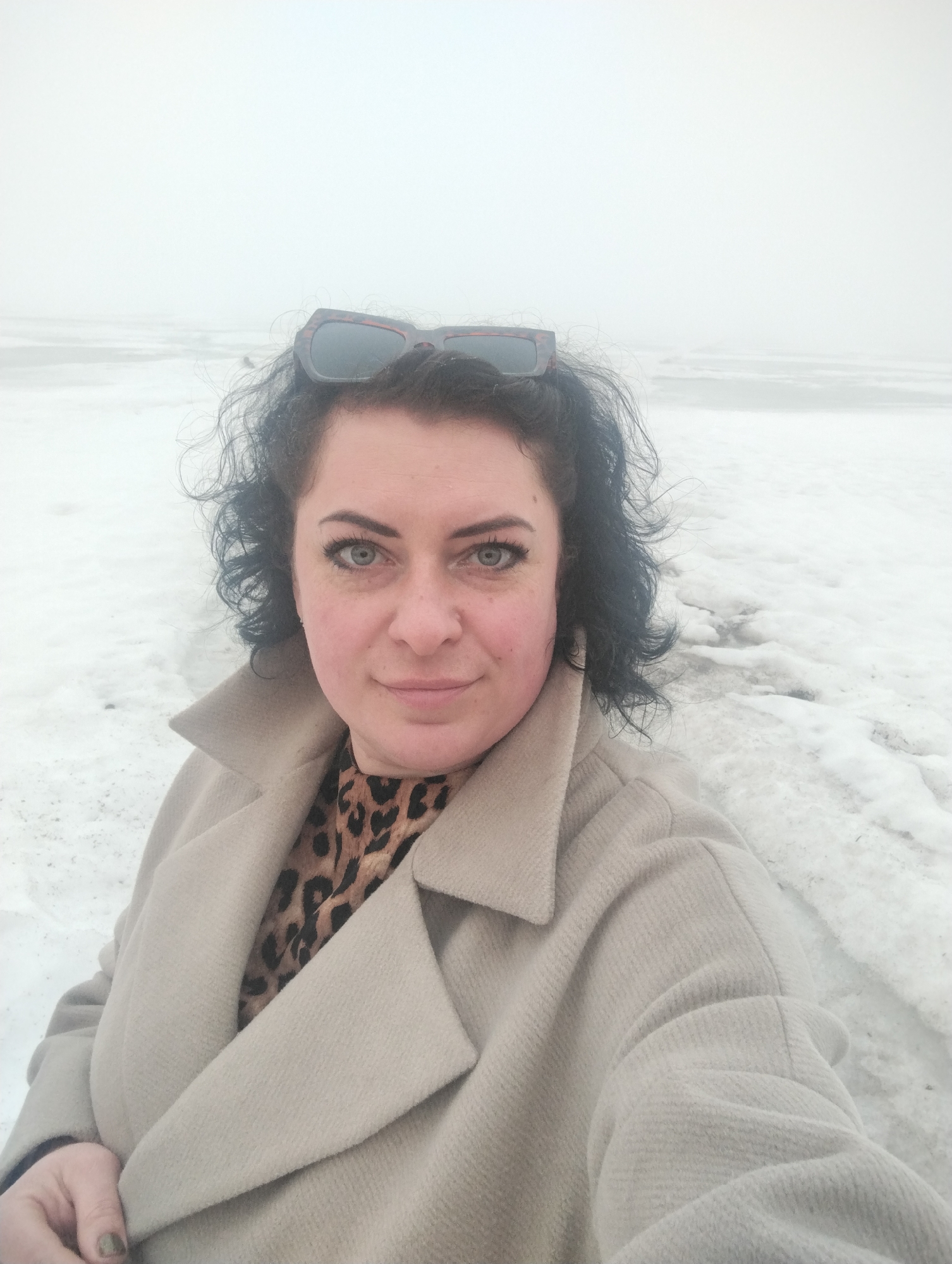 A selfie shows a woman in a light brown coat and leopard print shirt with sunglasses on top of her head. She is standing in an area covered with snow and ice. The background is overcast and obscured by fog or haze. The location is possibly Votkinsk, Russia, given the prompt, though there is no visual confirmation of the specific location in the image.