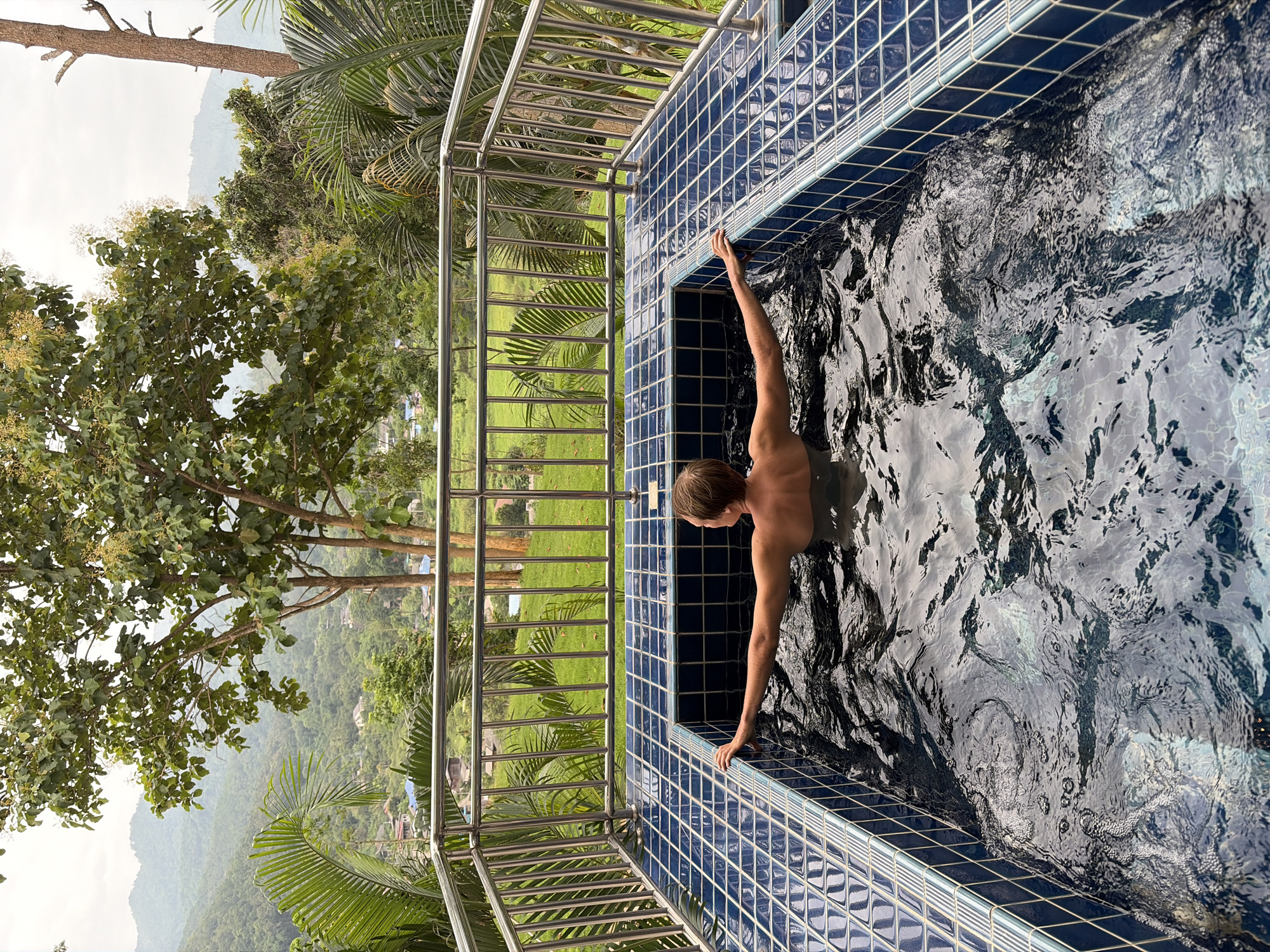 A person is relaxing in a hot tub overlooking a scenic view in Ban Kao Dua, Thailand. The hot tub is tiled with blue squares and has a metal railing around it. The person is facing away from the camera, with their arms resting on the edge of the hot tub. In the background, there is a lush green landscape with trees, mountains, and a village. The sky is overcast, suggesting a cloudy day.