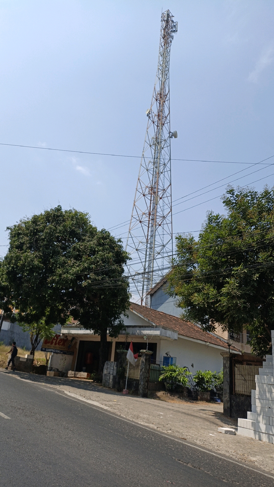 The image captures a view of a telecommunications tower against a clear blue sky, situated in Kuwasen, Indonesia. In the foreground, a paved road with a slight incline leads the eye towards a low-rise building with a tiled roof. Several trees with dense green foliage are visible on the left and right sides of the building, partially obscuring it. A person is seen walking on the sidewalk in front of the building. The building itself appears to be a commercial establishment, with a sign that reads "flex" and "TR. GAMPER SONO". A small Indonesian flag is planted in front of the building. The telecommunications tower, a tall lattice structure, dominates the background and is equipped with multiple antennas. Electrical wires are strung across the scene, further indicating an urban or semi-urban environment. The overall scene suggests a typical day in a residential or commercial area in Kuwasen, Indonesia.