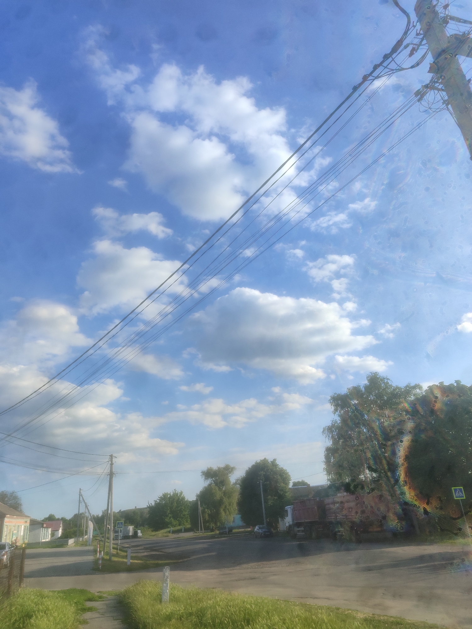 The image depicts a rural street scene under a partly cloudy blue sky. A power pole with numerous wires stretches diagonally across the frame from the upper right. In the foreground, there is a paved pathway bordered by tall green grass. The street curves to the right, with buildings visible on the left, including a light-colored structure with a green trim. On the right side of the street, several trees line the road. A large truck, appears to be a dump truck, is parked on the right side of the street. A pedestrian crossing sign is also visible on the right. The scene suggests a quiet, daytime atmosphere in a small town or village.