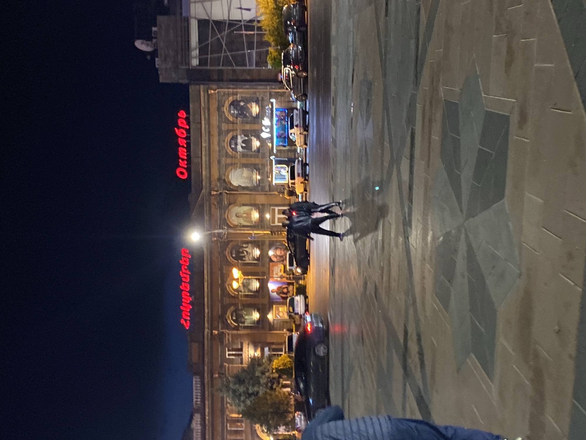 The image depicts the "October" cinema in Gyumri, Armenia, at night. The building facade is illuminated by red neon signs that spell out "Հոկտեմբեր" and "Октябрь". A dark-colored sedan is driving in the foreground, and two figures, likely pedestrians, are walking across the plaza in front of the cinema. Several other cars are parked or driving on the street adjacent to the building. The plaza features a decorative stone pattern on the ground, and streetlights illuminate the scene. The sky is dark, indicating it is nighttime.