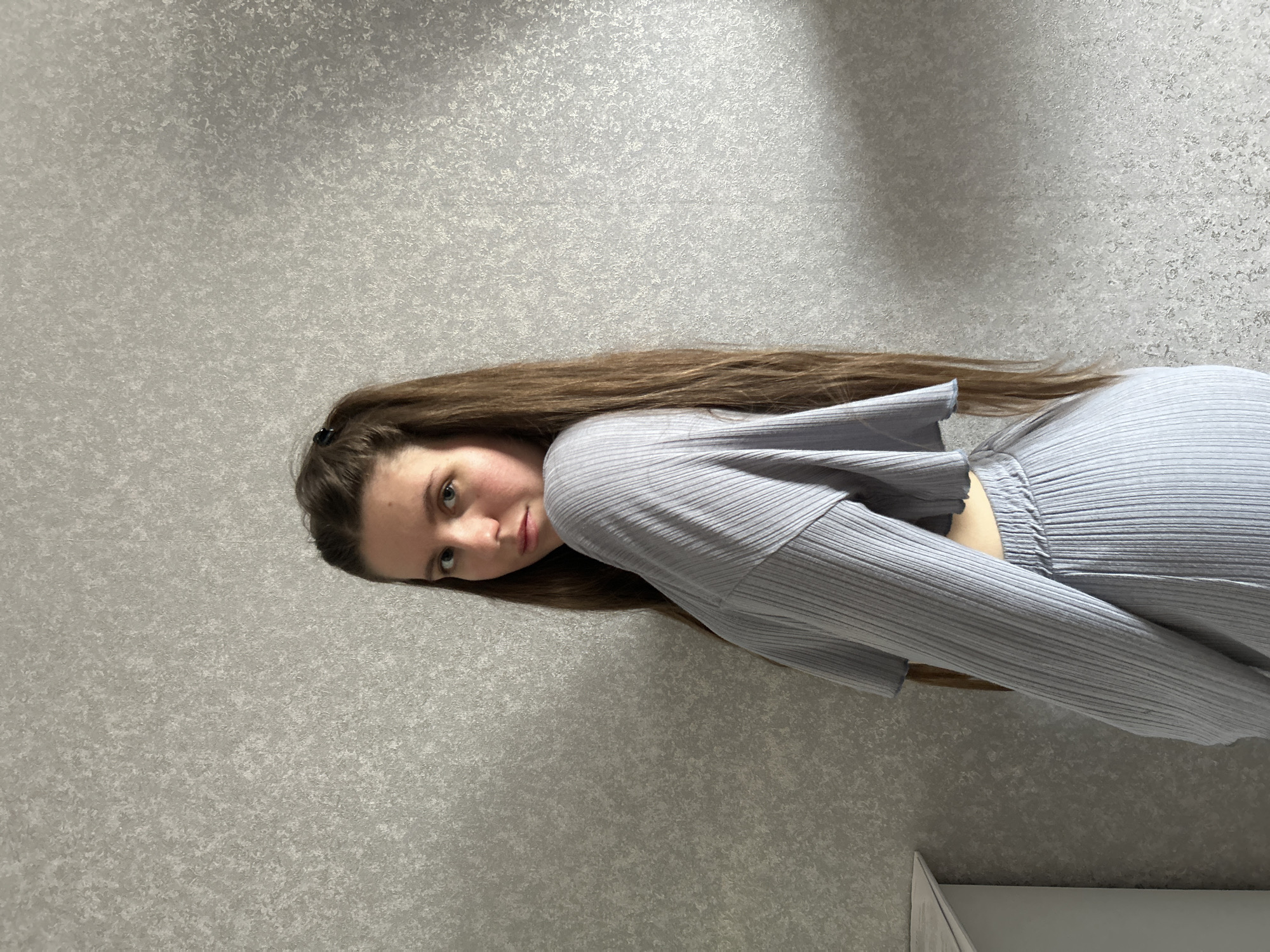 A woman is seen in a full-body shot, angled to the left. She has long brown hair. She is wearing a light blue ribbed two-piece outfit consisting of a top and pants. The background is a grey wall with a textured pattern. The bottom right corner shows a partial view of what appears to be a white cabinet. The location in the city of Klin, Russia, is not evident from the provided image.