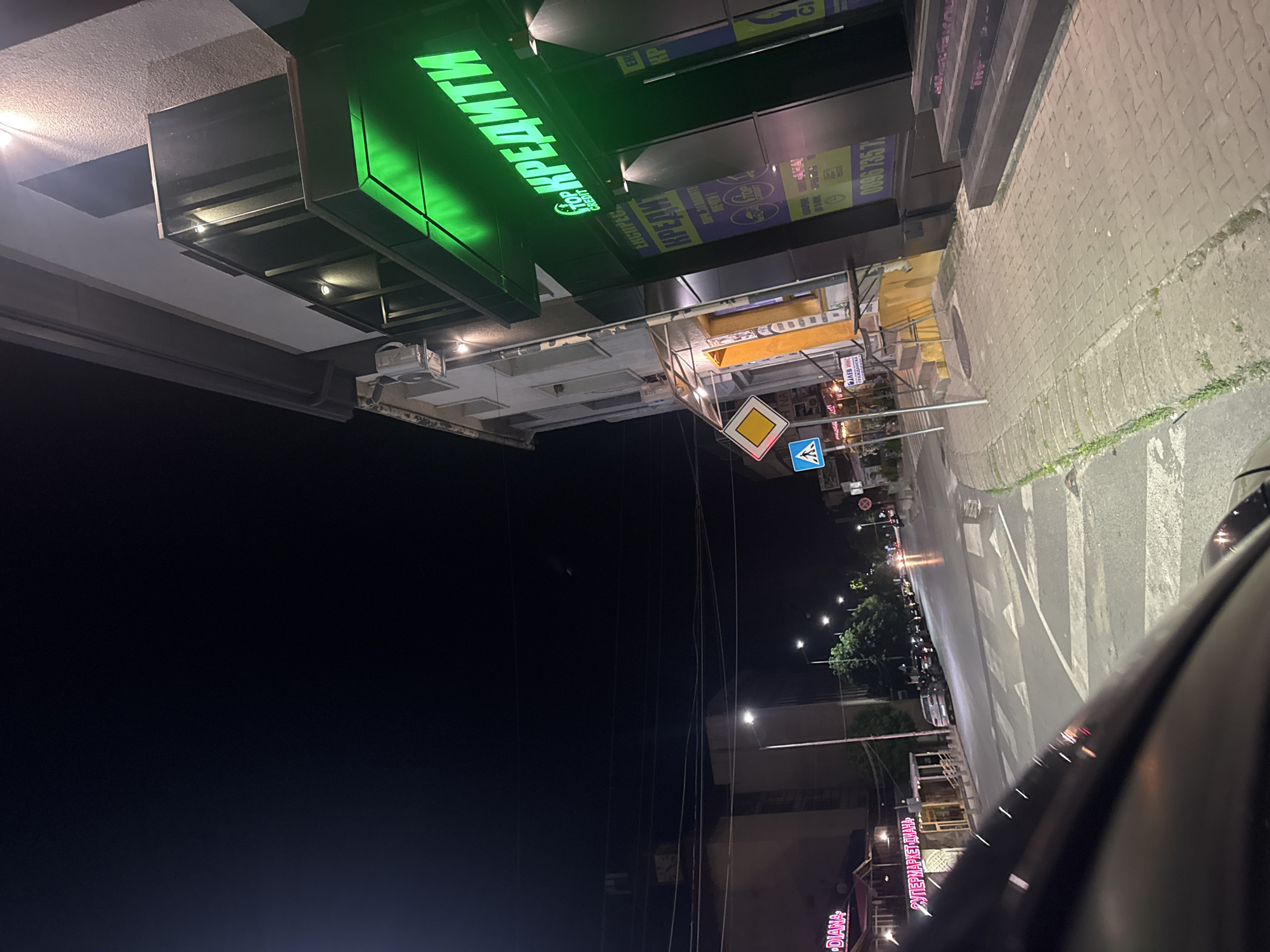 The image presents a nighttime scene from a street in an unknown city. A neon sign above a store, colored green and displaying the word "КРЕДИТИ" (CREDIT), dominates the top portion of the view. Various other signs and stores can be seen further down the street, including "DIANA" and "СУПЕРМАРКЕТ ДИАНА". The road is illuminated by streetlights, and there are pedestrian crossings. A car partially obscures the view from the right side of the image, suggesting the photograph was taken from within the vehicle. Additional street signs are present including a warning sign and one designating a pedestrian crossing.