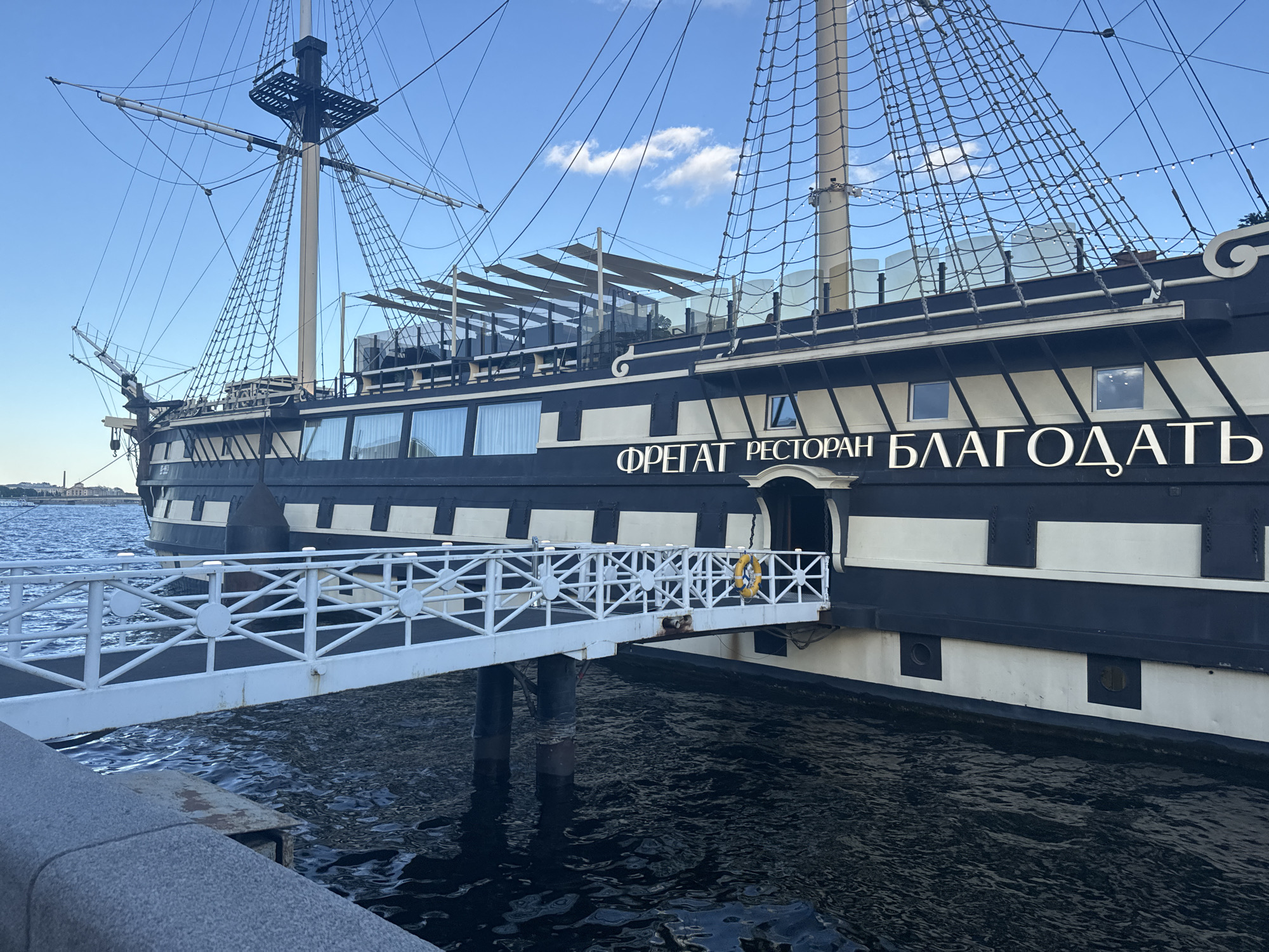 The image shows a black and white frigate restaurant named "Blagodat" docked on the Neva River in Saint Petersburg, Russia. A white gangway extends from the embankment to the ship's entrance. The ship has multiple decks, masts, and rigging, suggesting a historical design. The sky is blue with scattered clouds, and the water of the river is dark and rippling. There are no people visible in the image. The restaurant's name, written in Cyrillic characters, is prominently displayed on the side of the ship.