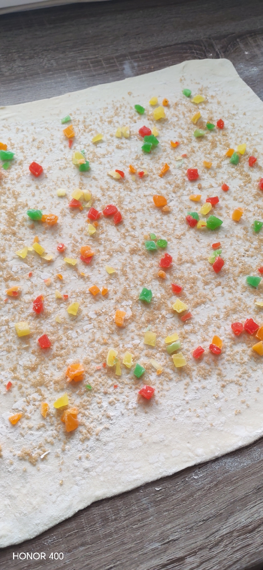 A rectangular slab of dough is spread out on a brown wooden surface. Scattered across the surface of the dough are small, colorful diced fruits (red, green, yellow, orange) and coarse brown sugar. A camera is placed in the lower left.