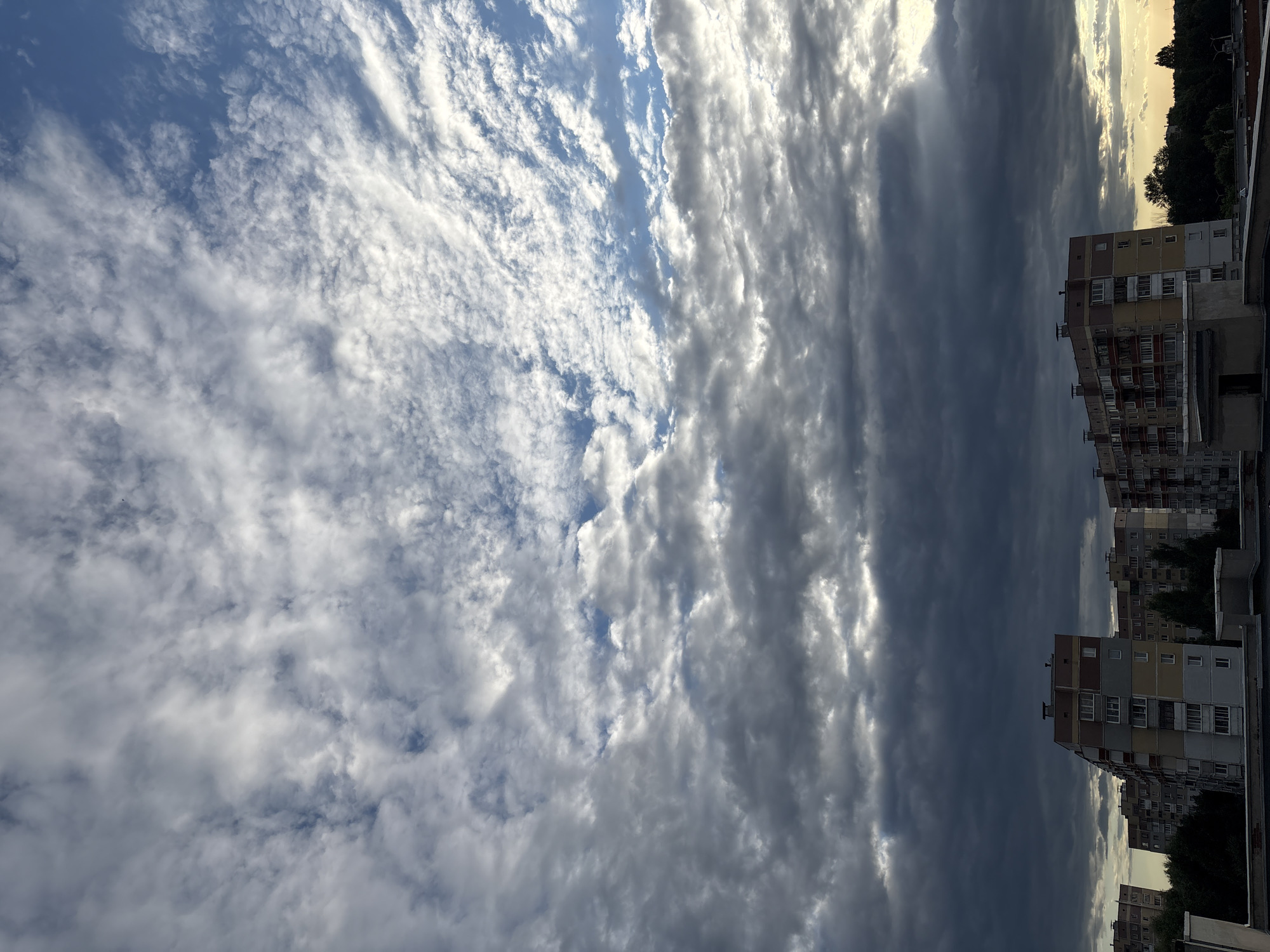 The image depicts a view of apartment buildings under a cloudy sky in the city of Vitsebsk, Belarus. The buildings are multi-story residential structures with balconies and windows, typical of Soviet-era architecture. Dark storm clouds dominate the upper portion of the frame, with some patches of lighter sky visible. The overall atmosphere suggests a pre-storm or post-storm condition. No people are visible in the scene. The environment appears to be urban, with the apartment blocks forming the primary man-made structures. Trees are visible in the background between the buildings. The lighting is diffused due to the cloud cover, with some highlights suggesting the sun is attempting to break through.