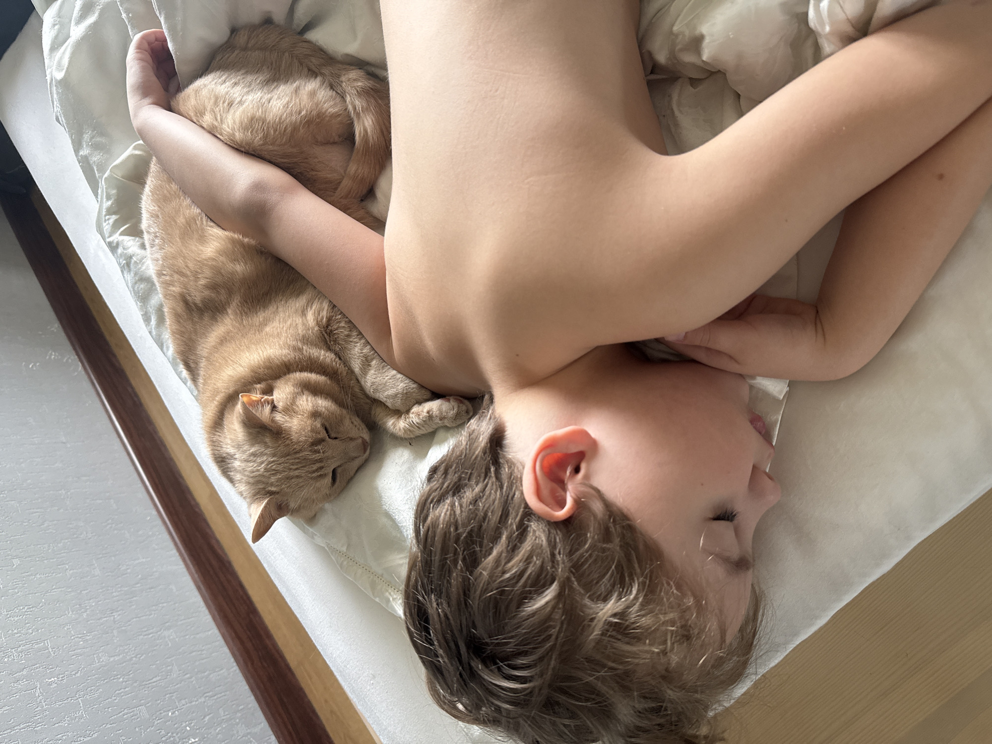 A young child and an orange tabby cat are lying on a white bed. The child is positioned with their head at the bottom of the frame, lying on their side. Their arms are stretched out above their head. The cat is lying next to the child, with their head near the top left corner of the frame. The scene is indoors, likely in a bedroom, as a wooden bed frame is visible. There is no information to determine this is in the city of Krasnoyarsk, Russia.