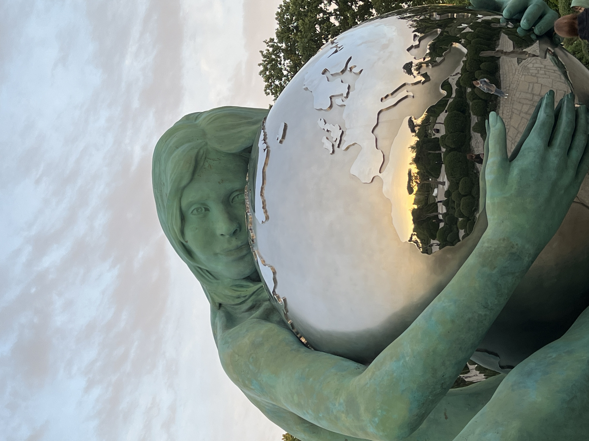 This vertical outdoor shot captures a large statue of a woman holding a reflective globe, likely situated in a public park or square in Krasnodar, Russia. The statue, made of metal, exhibits a distinctive greenish-blue patina. The woman's face is visible on the left, looking directly at the viewer with a serene expression, her sculpted hair framing her face. Her arms embrace the polished, metallic sphere.
The globe features a world map with continents, including Europe and parts of Asia, depicted as raised or cut-out elements. Its highly reflective surface mirrors the surroundings in intricate detail. A prominent golden reflection on the right side of the globe suggests the warm light of a setting or rising sun. Visible in the reflections are green trees, a paved ground area, and two individuals – one standing on the patterned ground and another partially seen at the top right, indicating other visitors are present.
The sky in the background is cloudy and bright, contrasting with the warm reflections on the globe. A dense green tree is also partially visible behind the globe. The perspective of the shot looks upwards towards the sculpture. No text is visible in the image.
