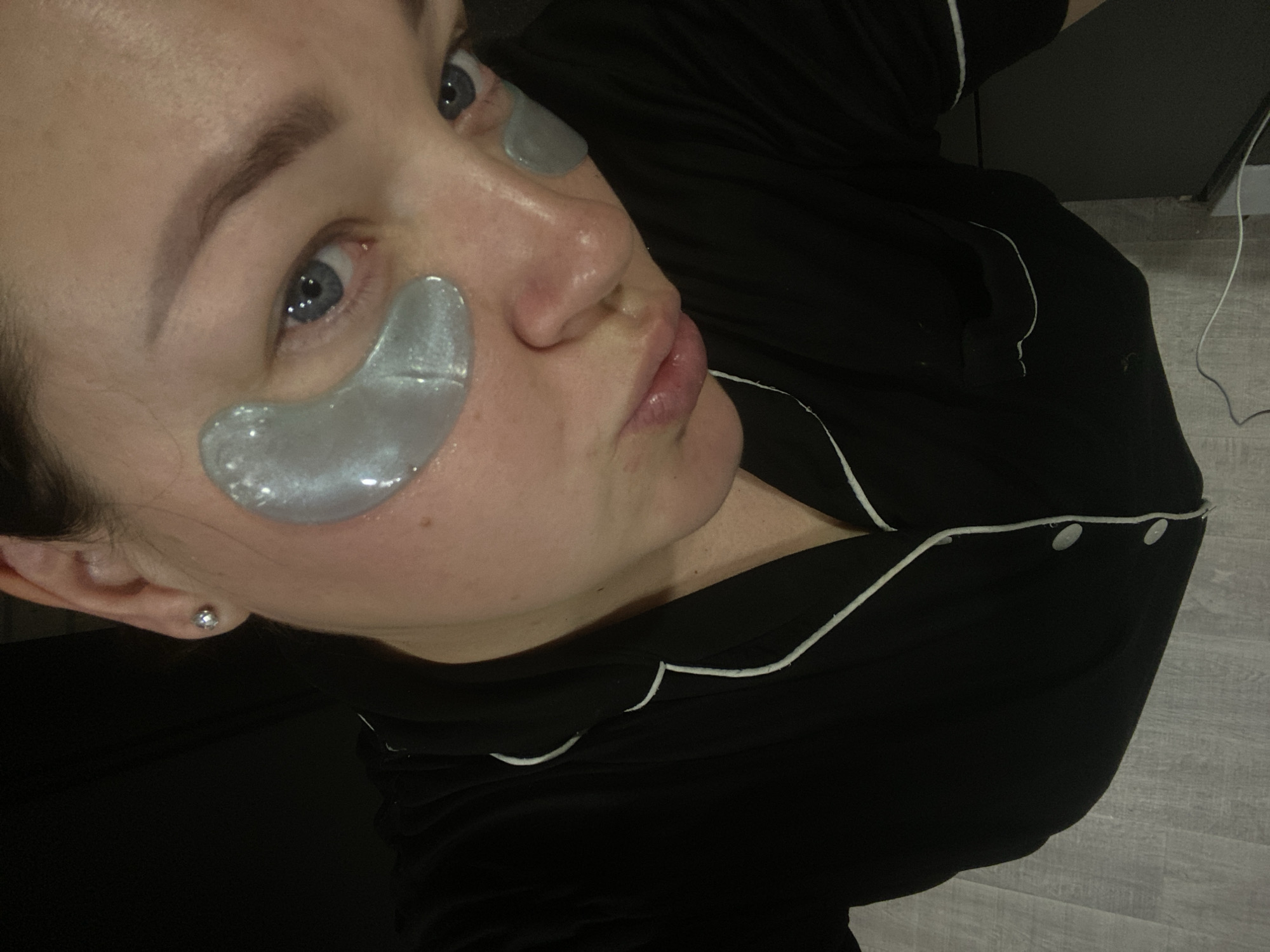 An individual is depicted in a close-up, high-angle indoor shot. The subject, an adult with medium-toned skin, is wearing a black collared top with white piping, consistent with pajamas or loungewear. Silver hydrogel under-eye patches are applied beneath each eye. A silver stud earring is visible in the right earlobe. The subject's hair is pulled back, and their blue eyes are directed towards the camera. Their lips are slightly pursed. The background immediately behind the subject is dark and indistinct. The lower portion of the image reveals a light grey floor with a plank-like pattern. A thin white cable lies on the floor towards the right side of the frame. The specific location within the city of Sochi, Russia, cannot be determined from the visual information provided in the image.